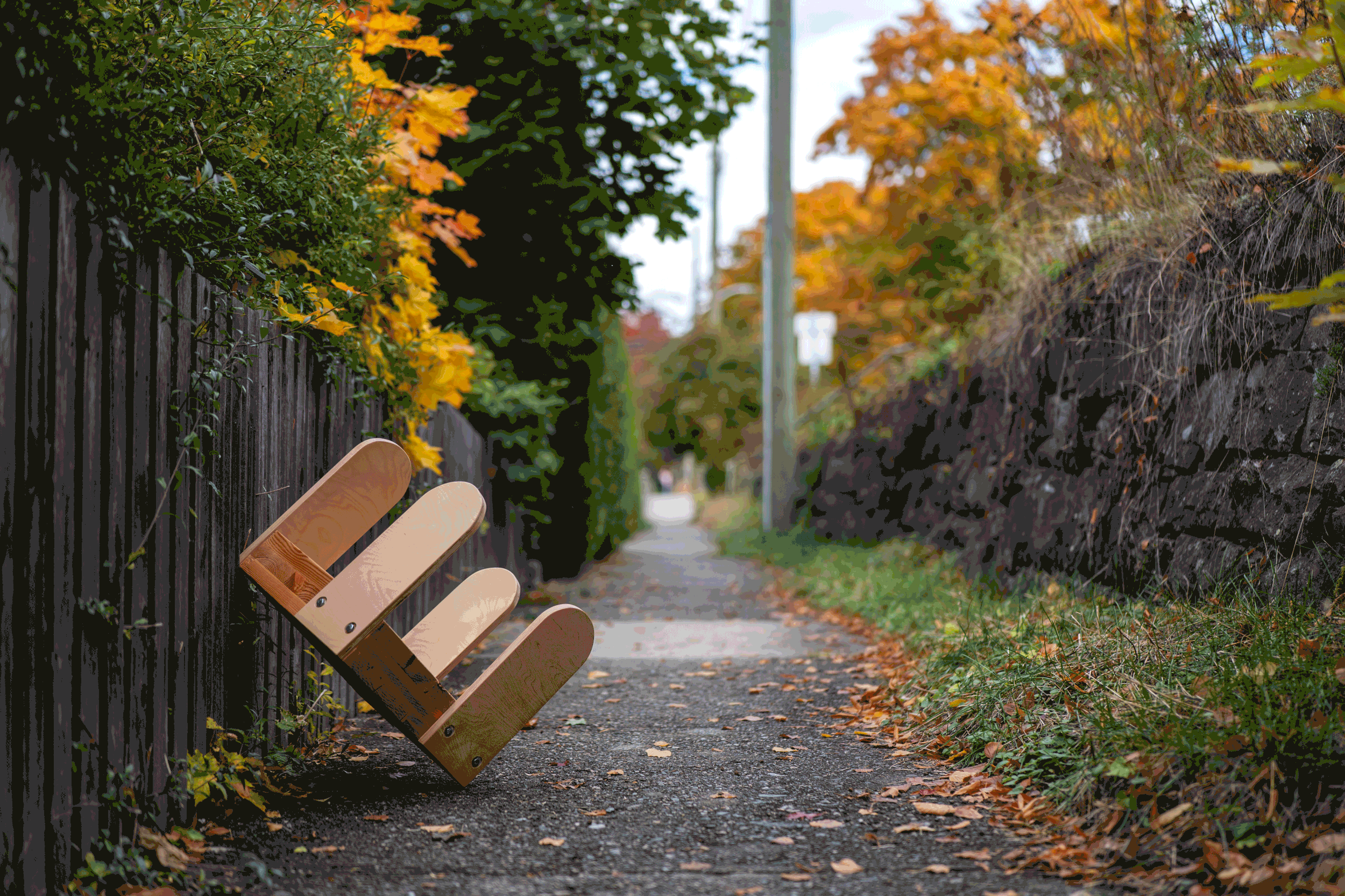 Empty wooden bench on a leaf-covered sidewalk in a suburban neighborhood during fall, with colorful trees and a stone wall on one side and a wooden fence on the other.