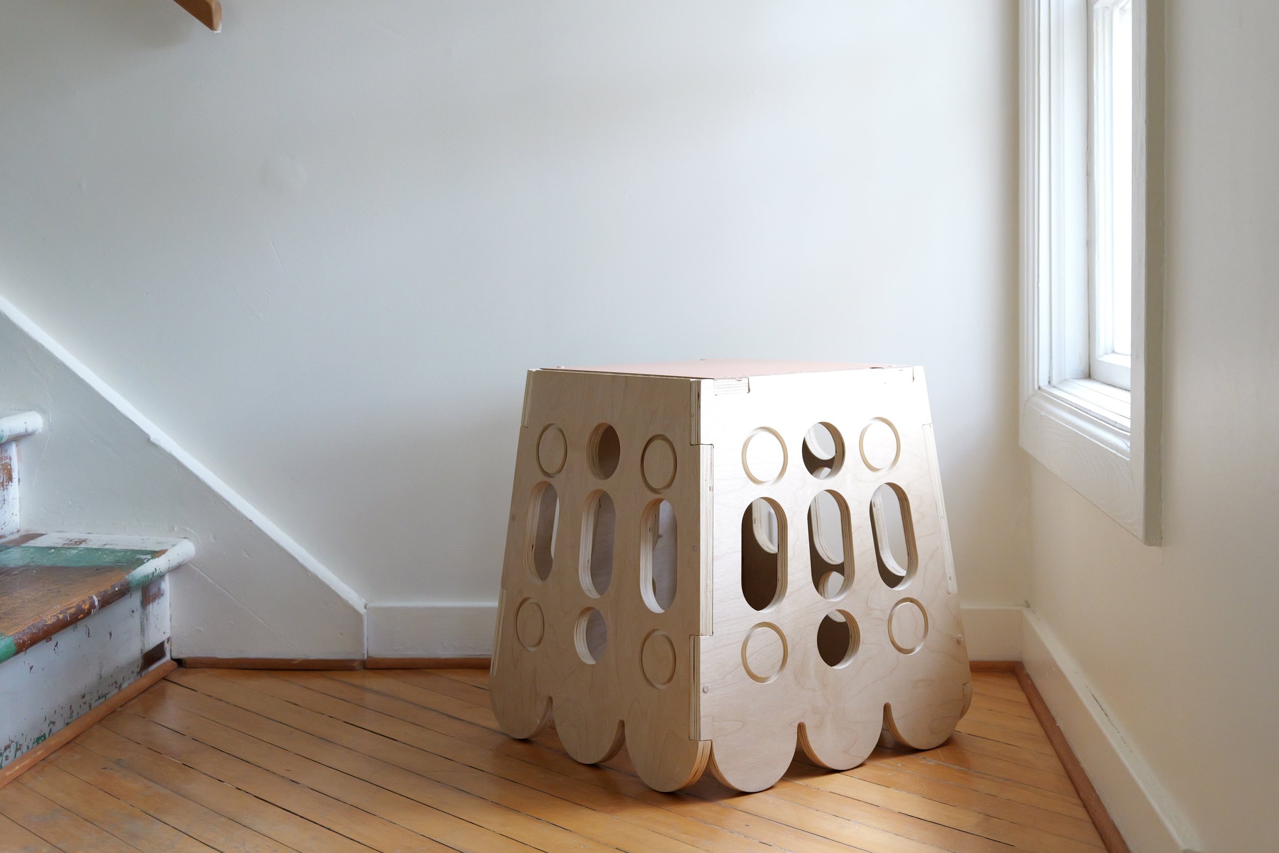 A wooden stool with circular and oval cutouts, placed on a hardwood floor near a staircase and a window in a room with white walls.
