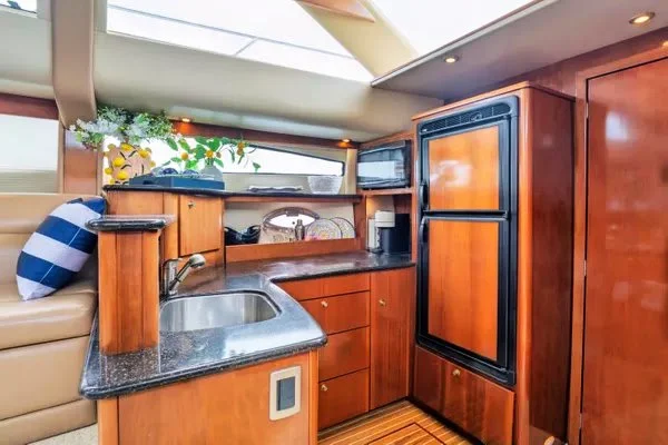 Compact kitchen area on a boat with wooden cabinetry, a stainless steel sink, a black refrigerator, and a small microwave, with a window above and decorative plants on the windowsill.