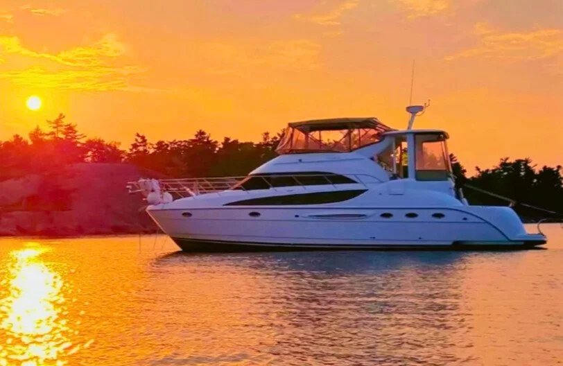 A white yacht floating on calm water during a sunset with orange and yellow sky and silhouettes of trees in the background.