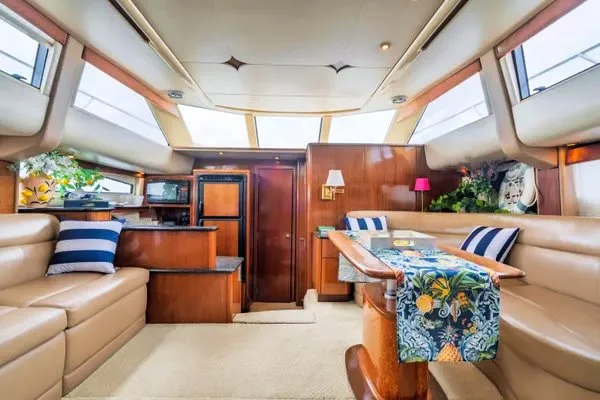 Interior of a boat or yacht with beige couches, colorful pillows, a wooden table with a tablecloth, and large windows with sunny views.