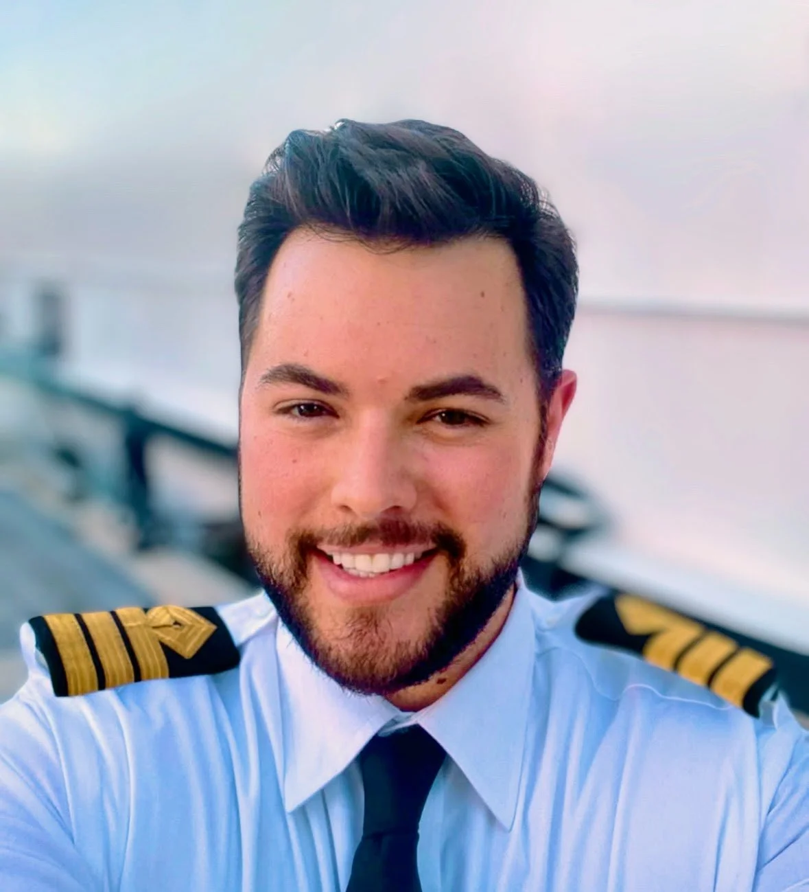 A smiling man in airline pilot uniform with epaulets on shoulders, taking a selfie.