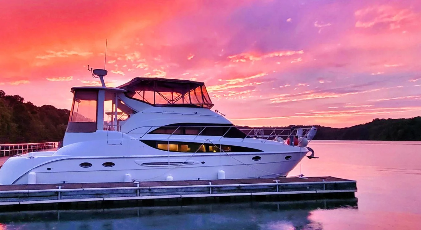A white yacht docked at a pier during a colorful sunset with pink and purple skies and water reflections.