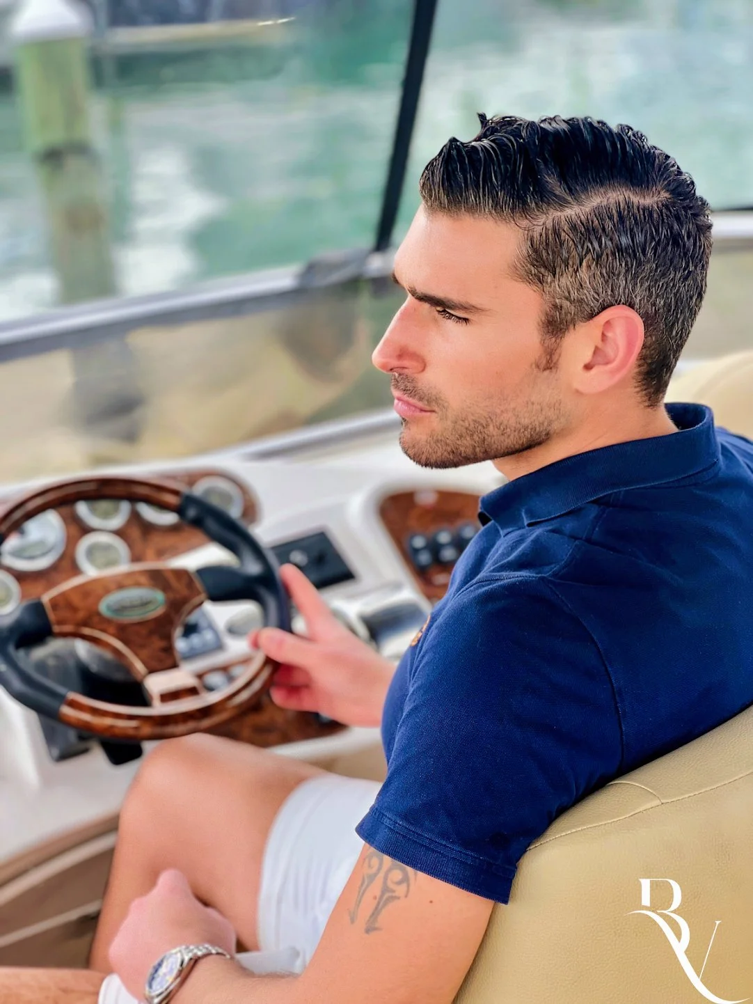 A man with dark hair and a tattoo on his arm sitting in the driver's seat of a boat, wearing a blue shirt and light-colored shorts, holding the steering wheel and looking ahead, with water and part of the boat visible outside the window.