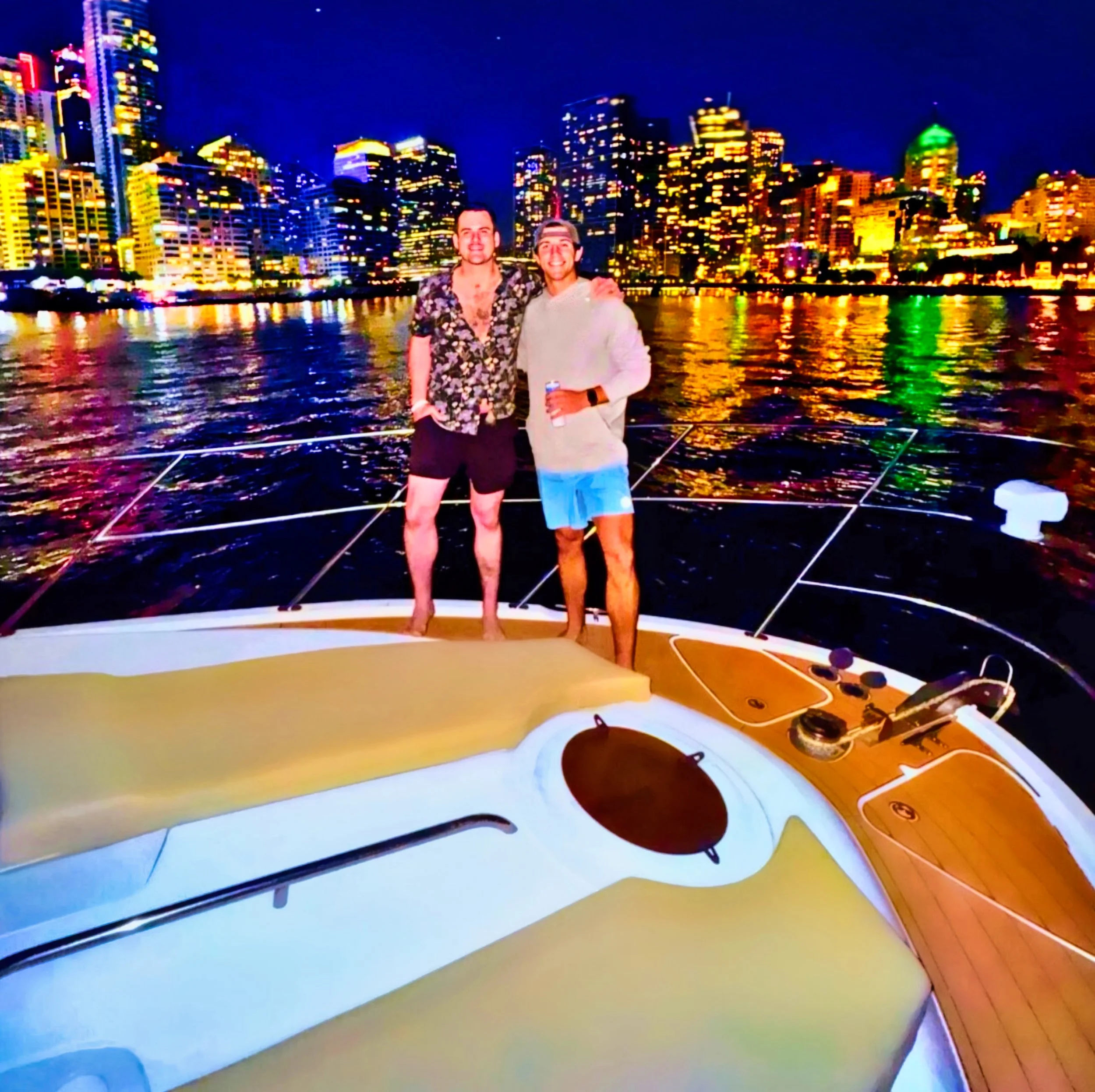 Two men standing on a boat at night with a city skyline and colorful lights reflecting on the water behind them.