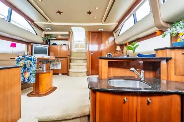 Interior of a boat or yacht with wooden cabinets, a small kitchen area with a sink, a TV, a dining table with a floral chair, and windows with blinds.