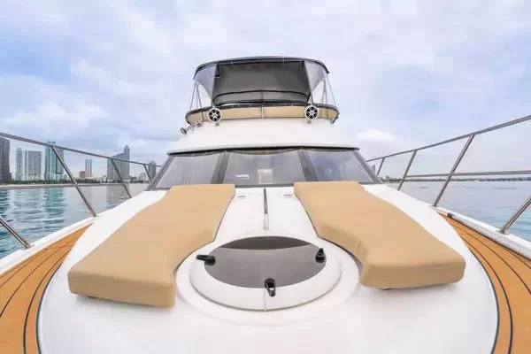Front view of a white yacht with two beige sunbathing cushions, a circular hatch, and city skyline in the background.