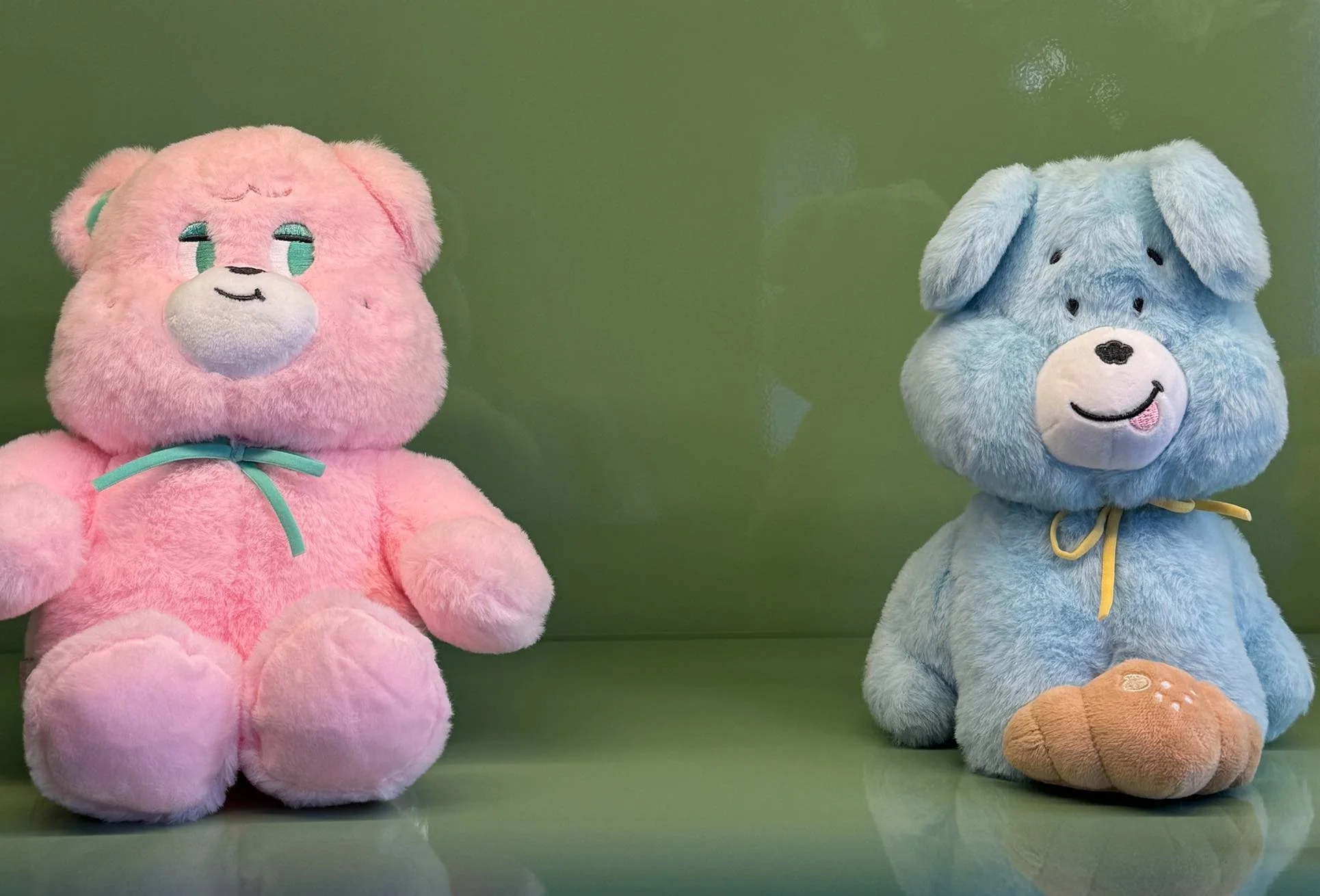 Two plush stuffed animals, a pink bear on the left and a blue dog on the right, sitting on a shiny green surface against a light green background.
