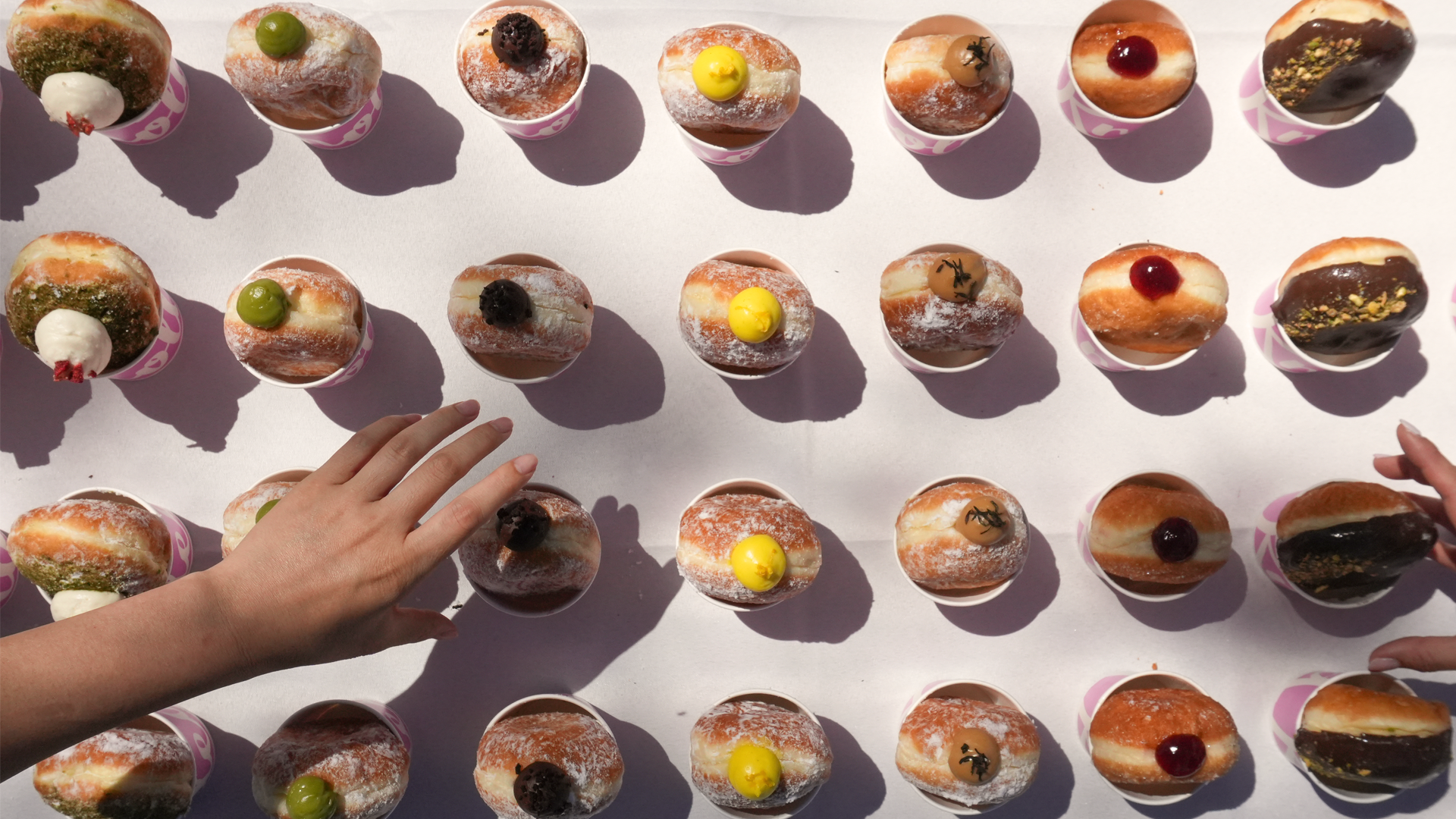 Top-down view of rows of cupcakes with frosting and colorful toppings on a white surface, with two hands reaching towards the cupcakes.