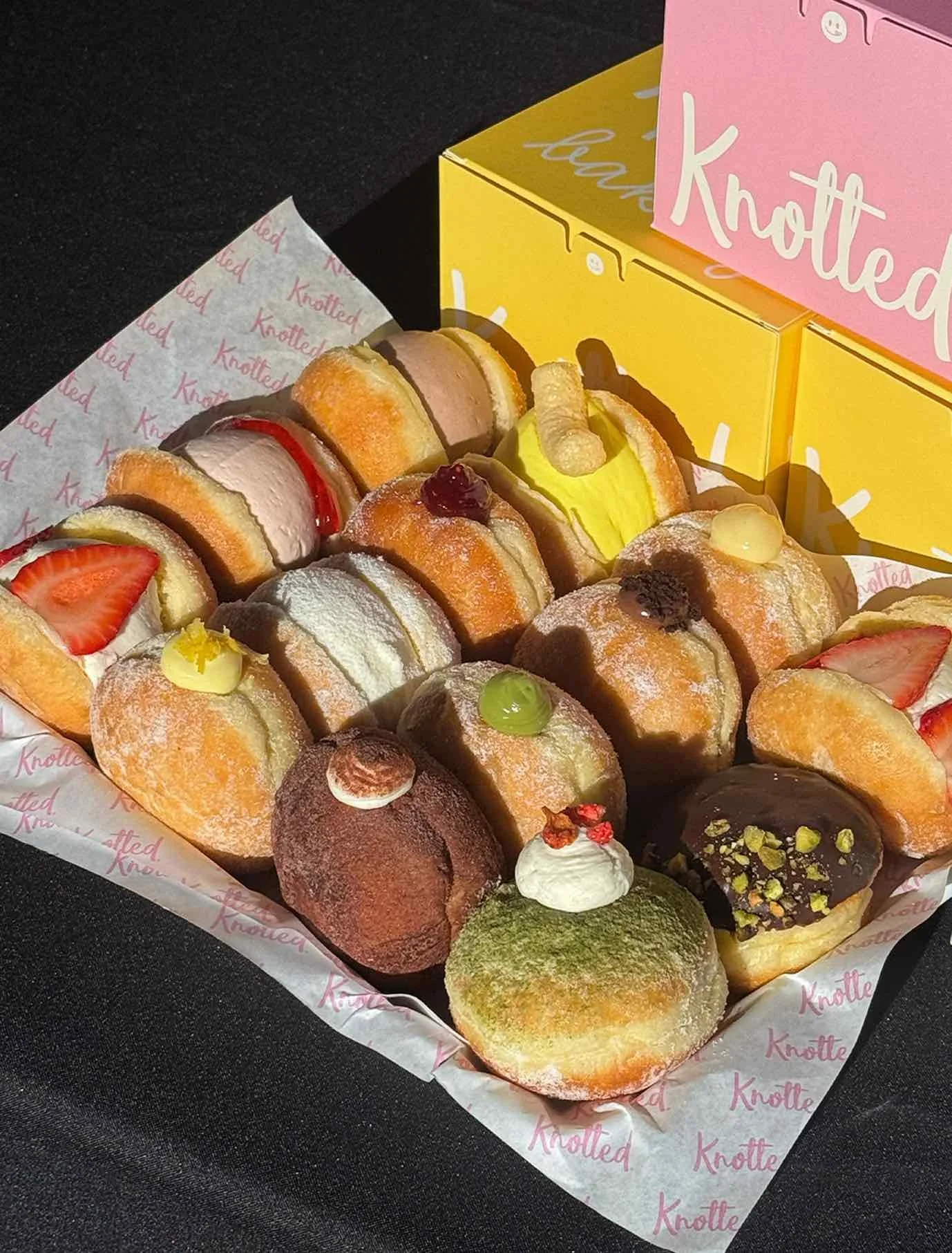 Assorted filled donuts with strawberries, cream, and toppings arranged in a Knotted-branded catering box.