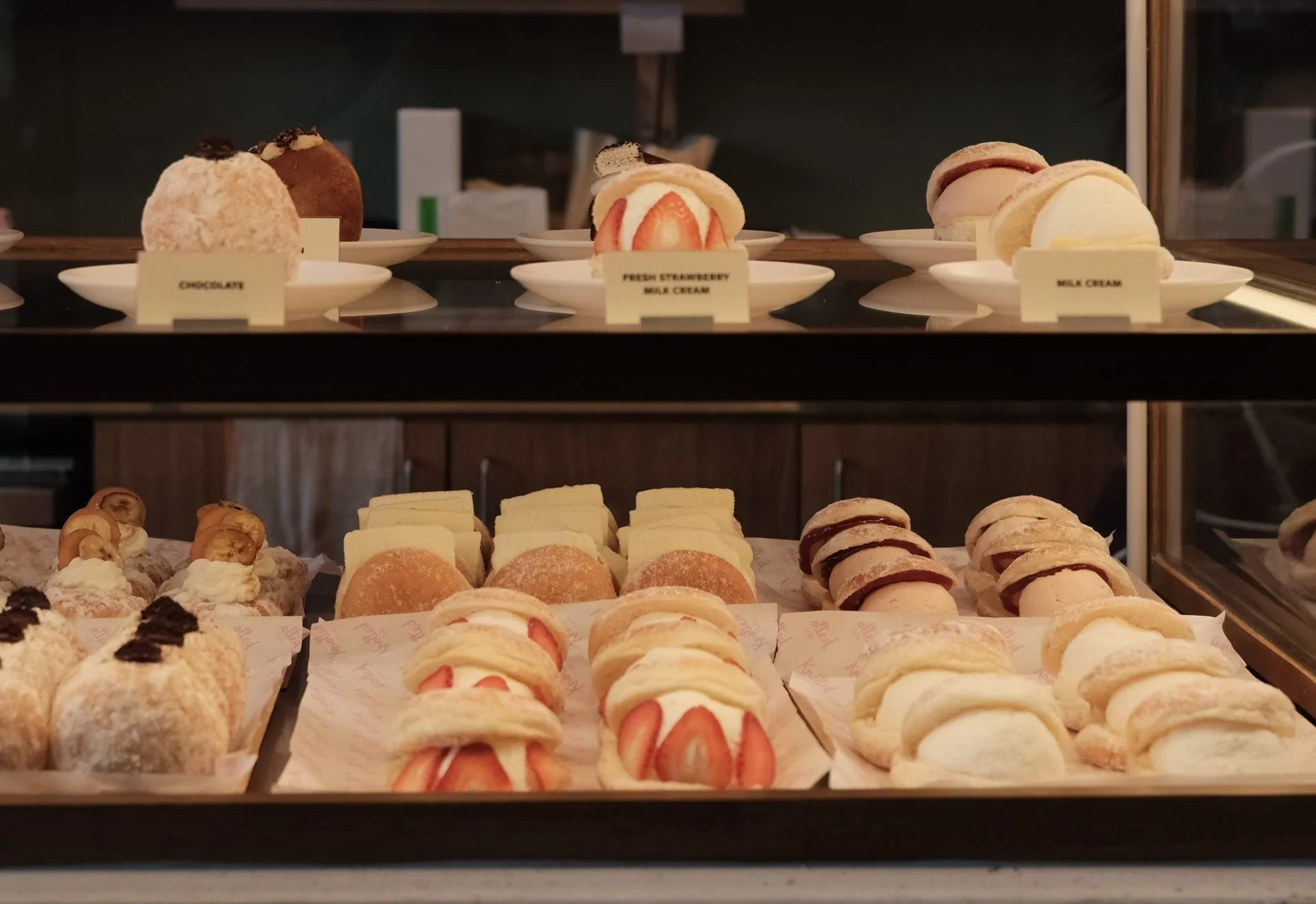 Display case with assorted desserts, including strawberry and cream pastries, macarons, and cake slices, with labels for chocolate, strawberry milk cream, and milk cream.