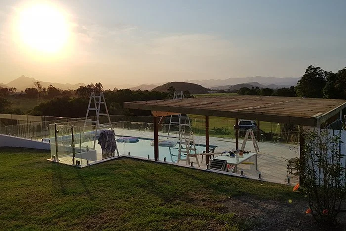 A backyard swimming pool with a wooden pergola under construction, ladders, and tools around. The sun is setting in the background over distant hills and trees.