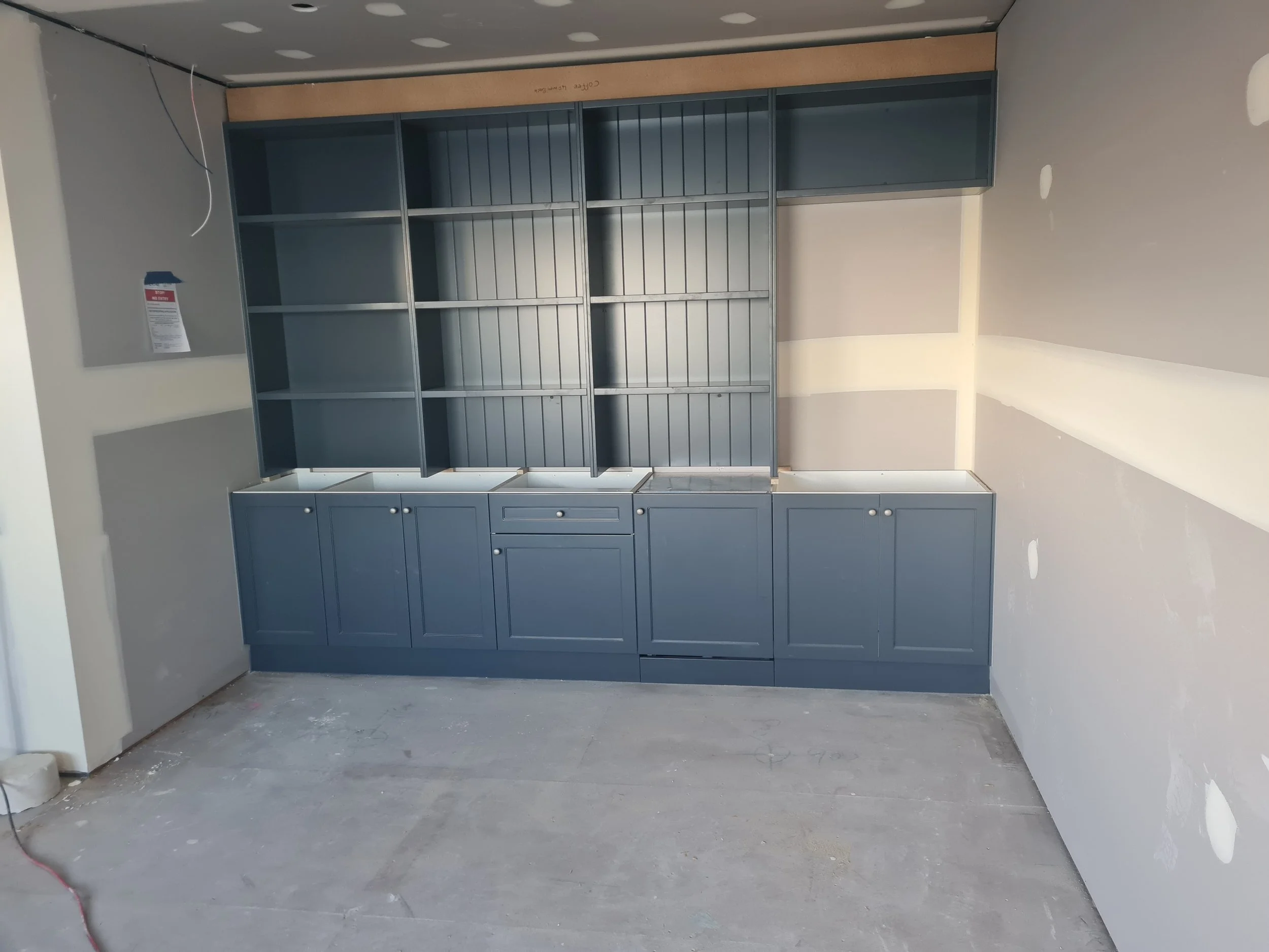 Unfinished interior with blue cabinets and shelving against a partly constructed wall, exposed wiring above, and bare concrete floor.