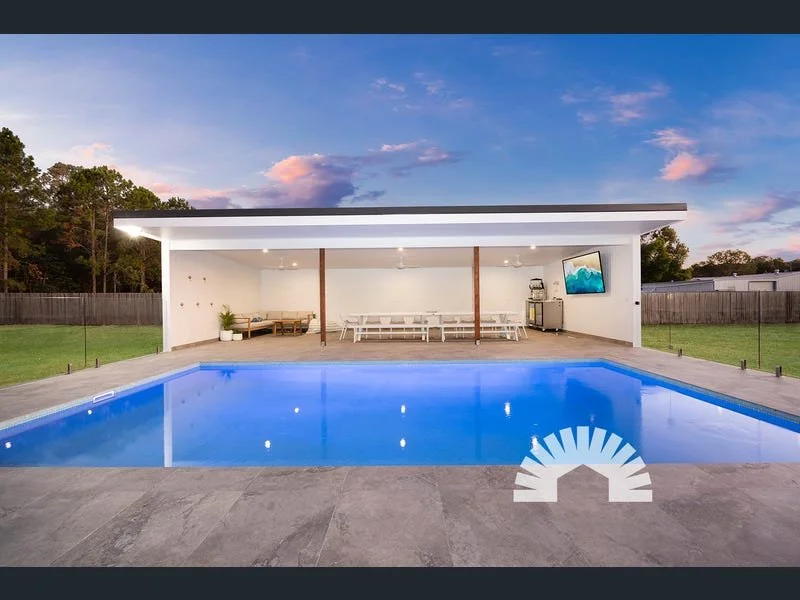 Modern backyard with swimming pool, covered patio area with seating and TV, surrounded by grass and trees, during sunset.