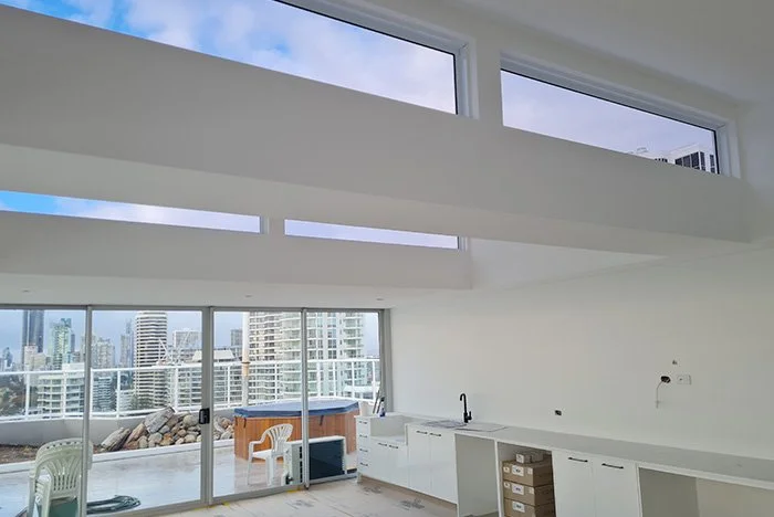 Modern apartment interior with large windows offering city views, unfurnished room with white kitchen cabinets, sliding glass doors, and balcony with outdoor chairs in Northern NSW Australia