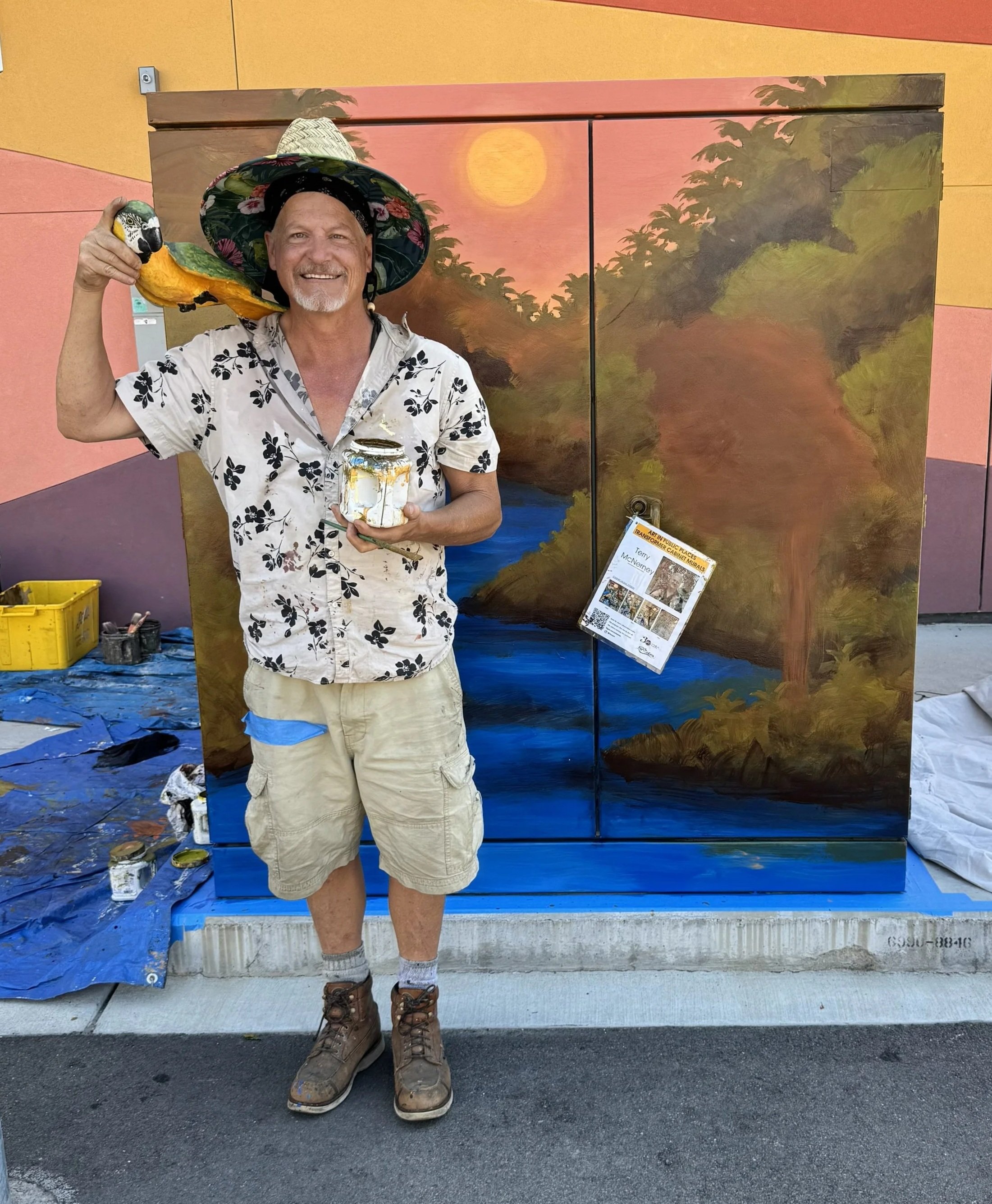 Terry McNerney with a parrot on his shoulder, holding a jar, standing in front of a painted mural of a river landscape with trees and a setting sun.