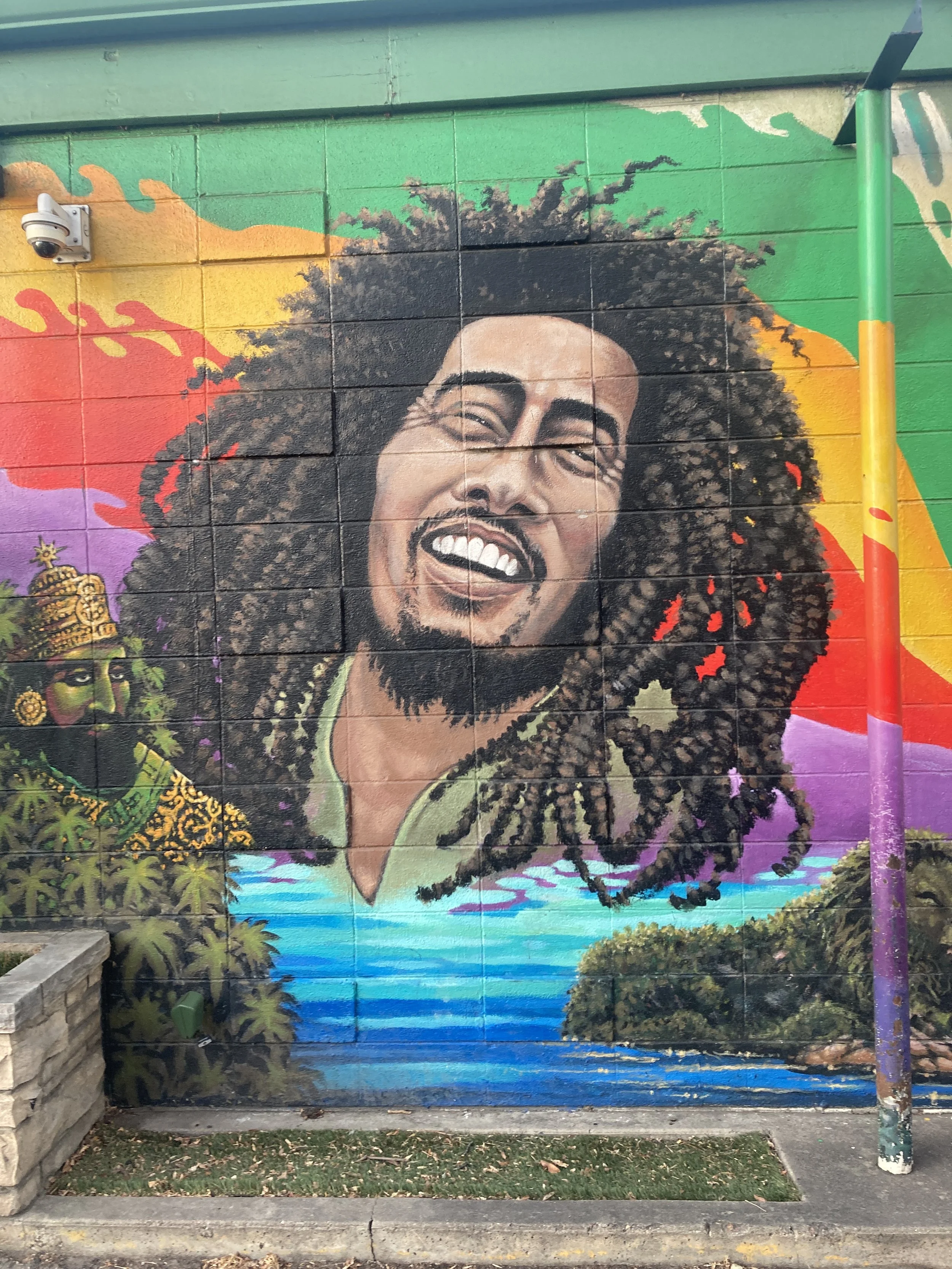 Colorful mural of Bob Marley with dreadlocks smiling, with tropical scenery including water, islands, and another person wearing a crown and jewelry in the background.