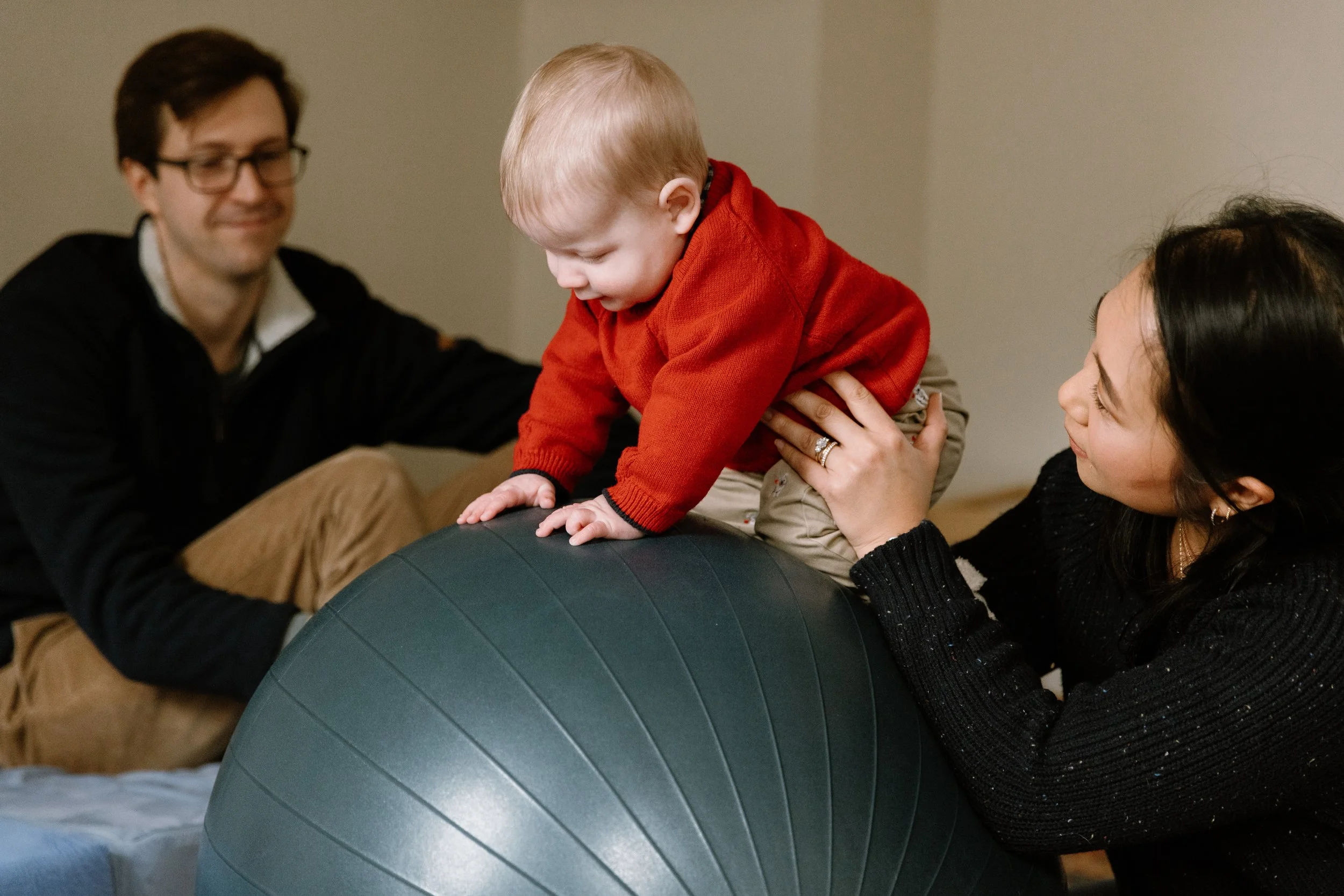 Baby in red sweater climbing on a balance ball supported by a woman, with a man watching.