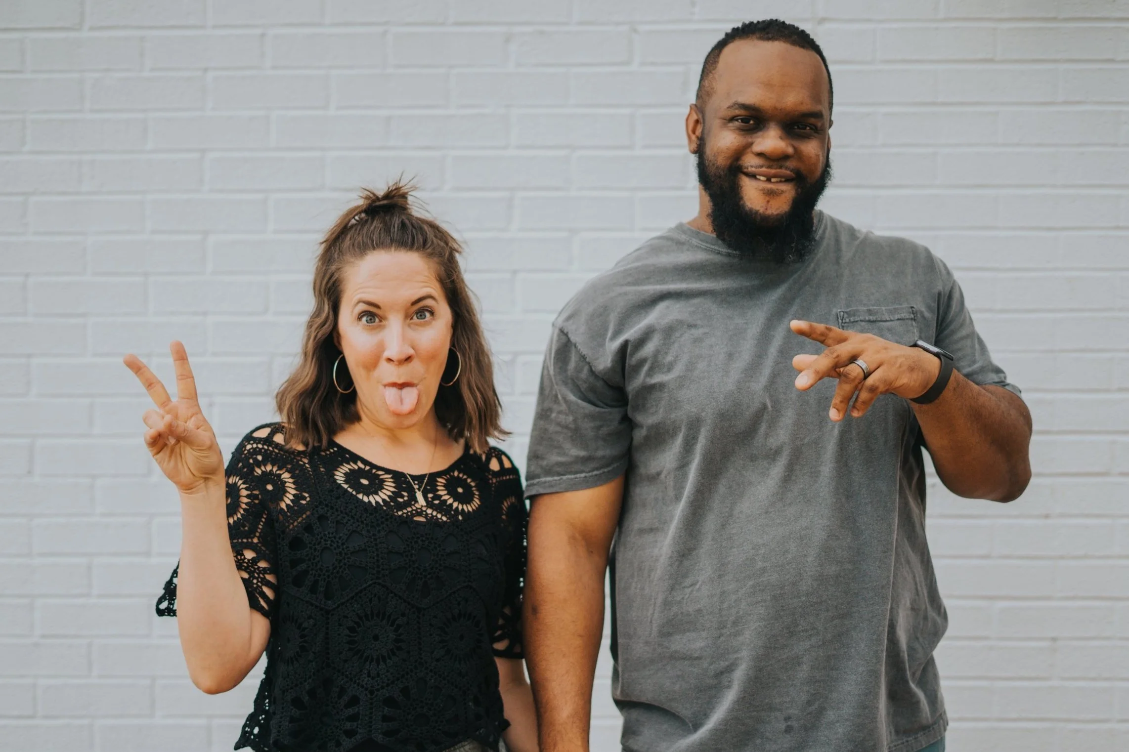 Two people making playful gestures in front of a brick wall, one with a peace sign and tongue out, the other with fingers pointed.