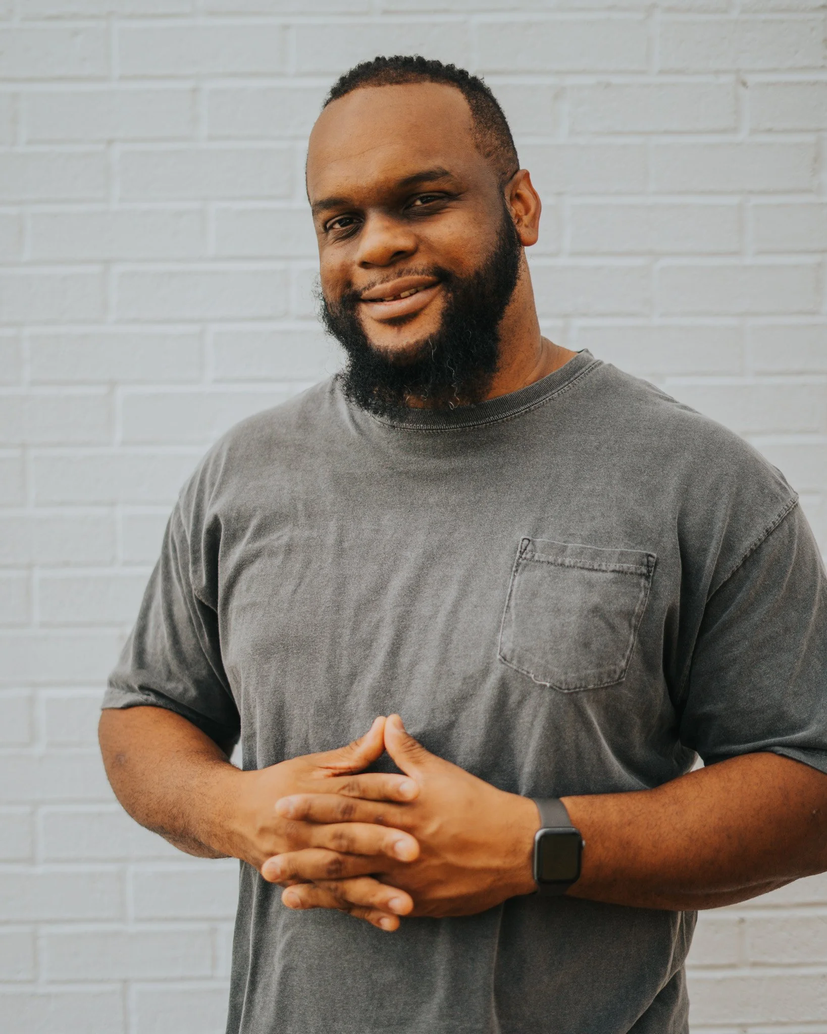 Man smiling, wearing a gray t-shirt with a watch, standing against a white brick wall.