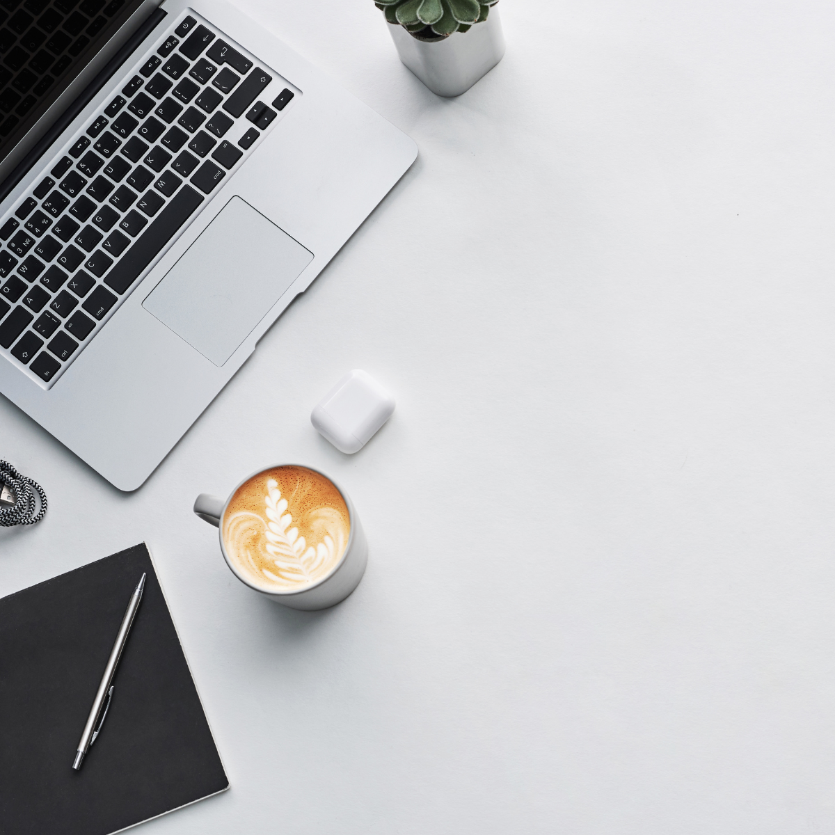 Minimalist workspace with laptop, coffee cup with latte art, notebook, pen, small plant, and wireless earbuds on a white desk.