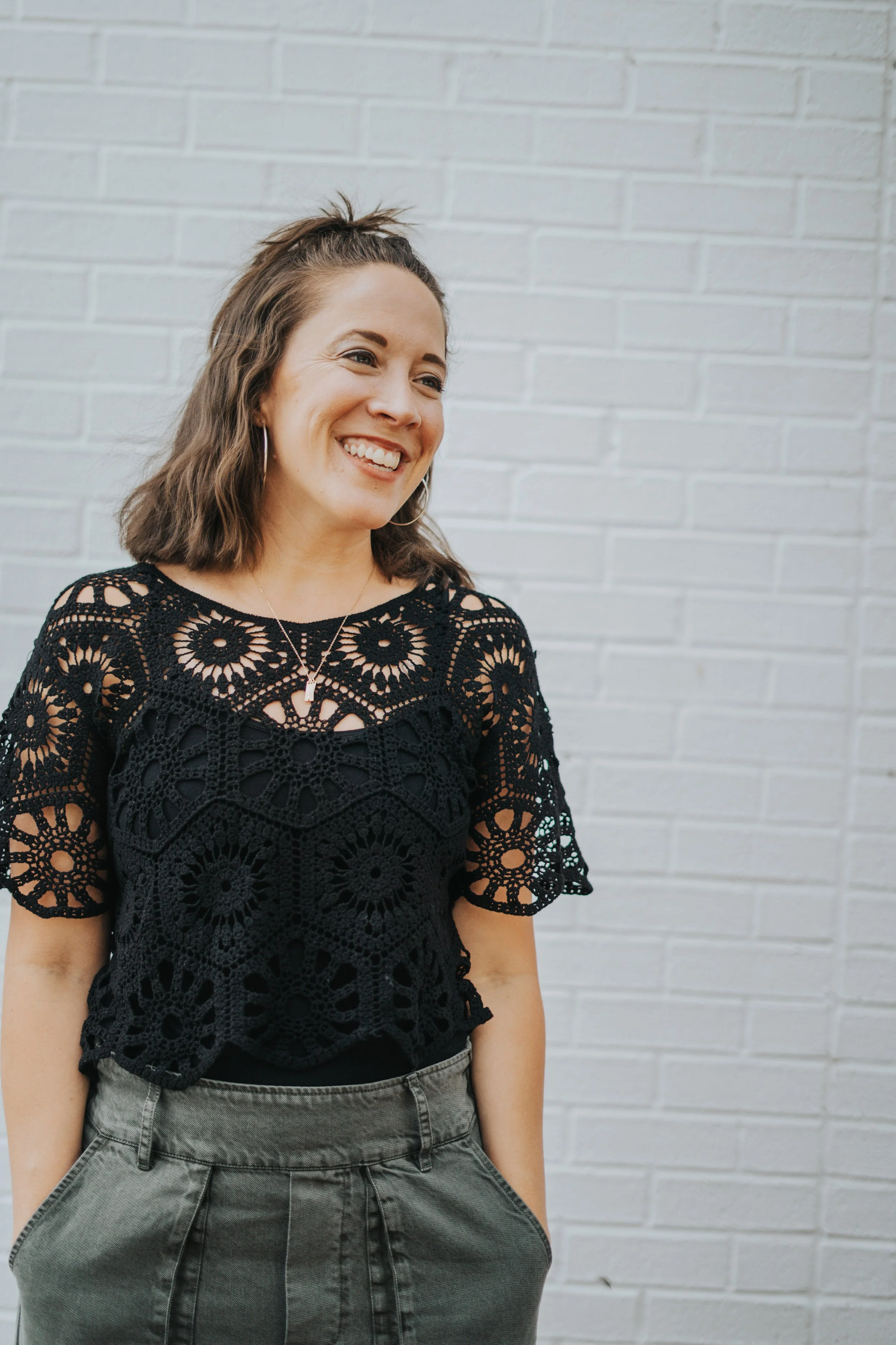 Woman wearing a black lace top smiles in front of a white brick wall.