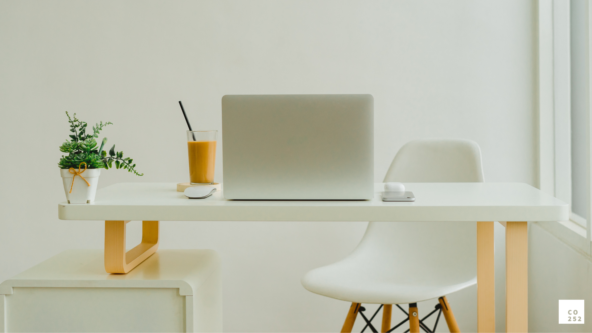 Minimalist workspace with laptop, orange drink, and potted plant on white desk, next to modern chair.