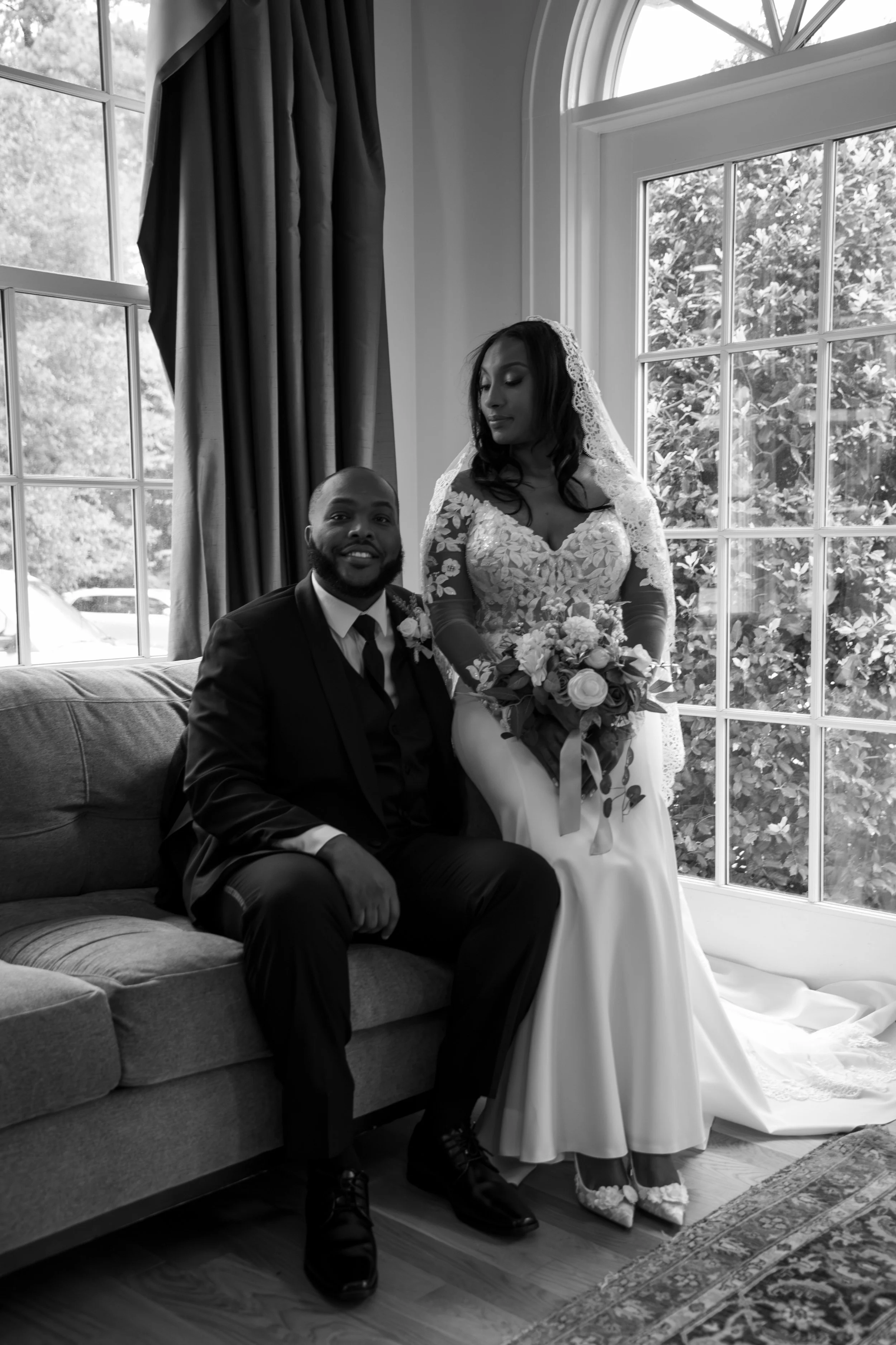 Photo & Video by Will Locke Wedding Photography at the Woman's Club of Portsmouth in Virginia. A bride in a white wedding dress holding a bouquet stands next to a groom seated on a sofa indoors near large windows with curtains.