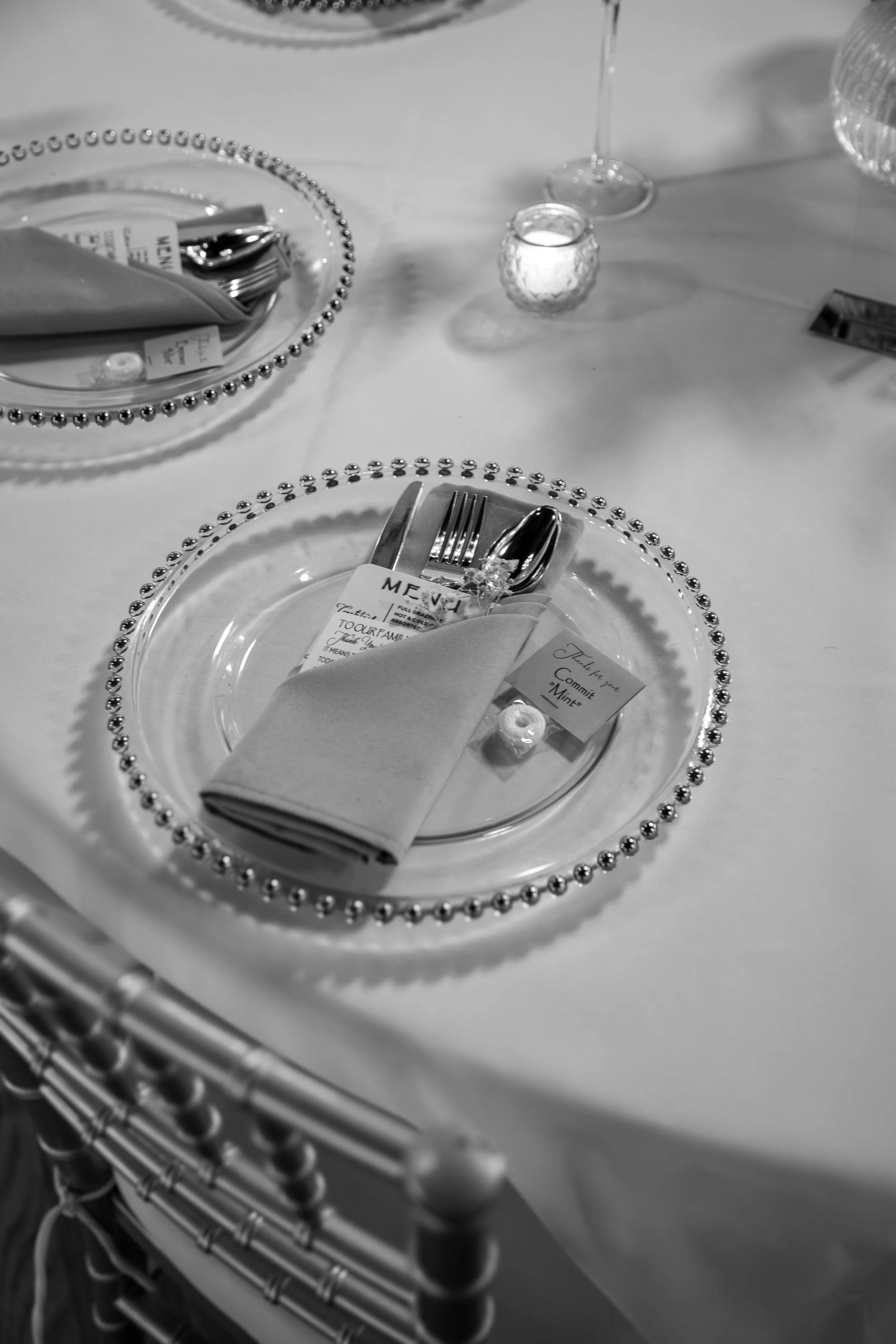 Wedding Photography by Will Locke. Elegant table setting with a plate bordered by metallic beads, silverware wrapped in a cloth napkin, a small dish with a pearl-like decoration, a menu, a small card, a glass of wine, and a lit candle.