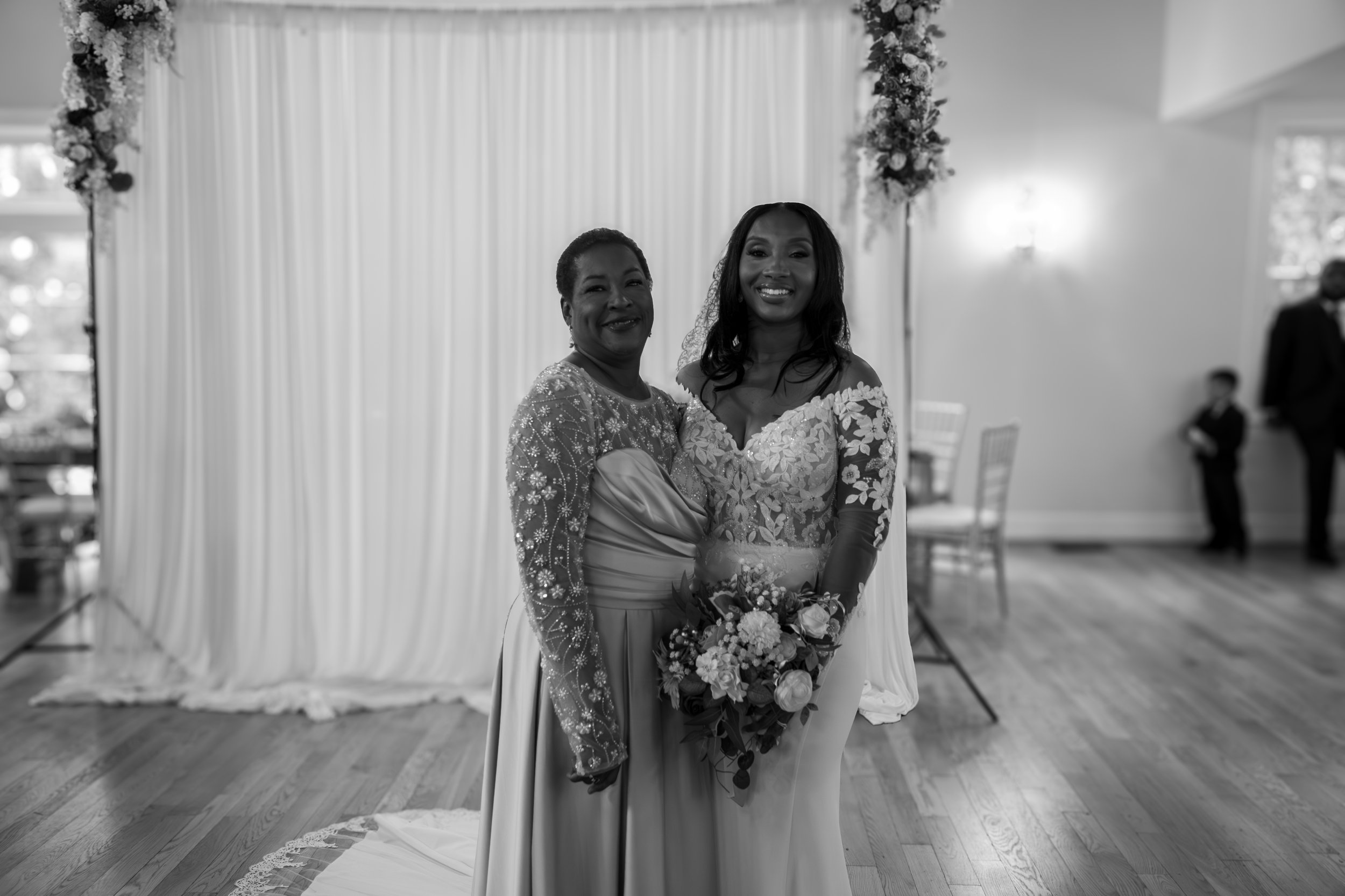 Photo & Video by Will Locke Wedding Photography at the Woman's Club of Portsmouth in VA. A bride in a lace wedding dress holding a bouquet, standing next to an older woman in a fancy dress, both smiling at the camera in a decorated wedding venue.