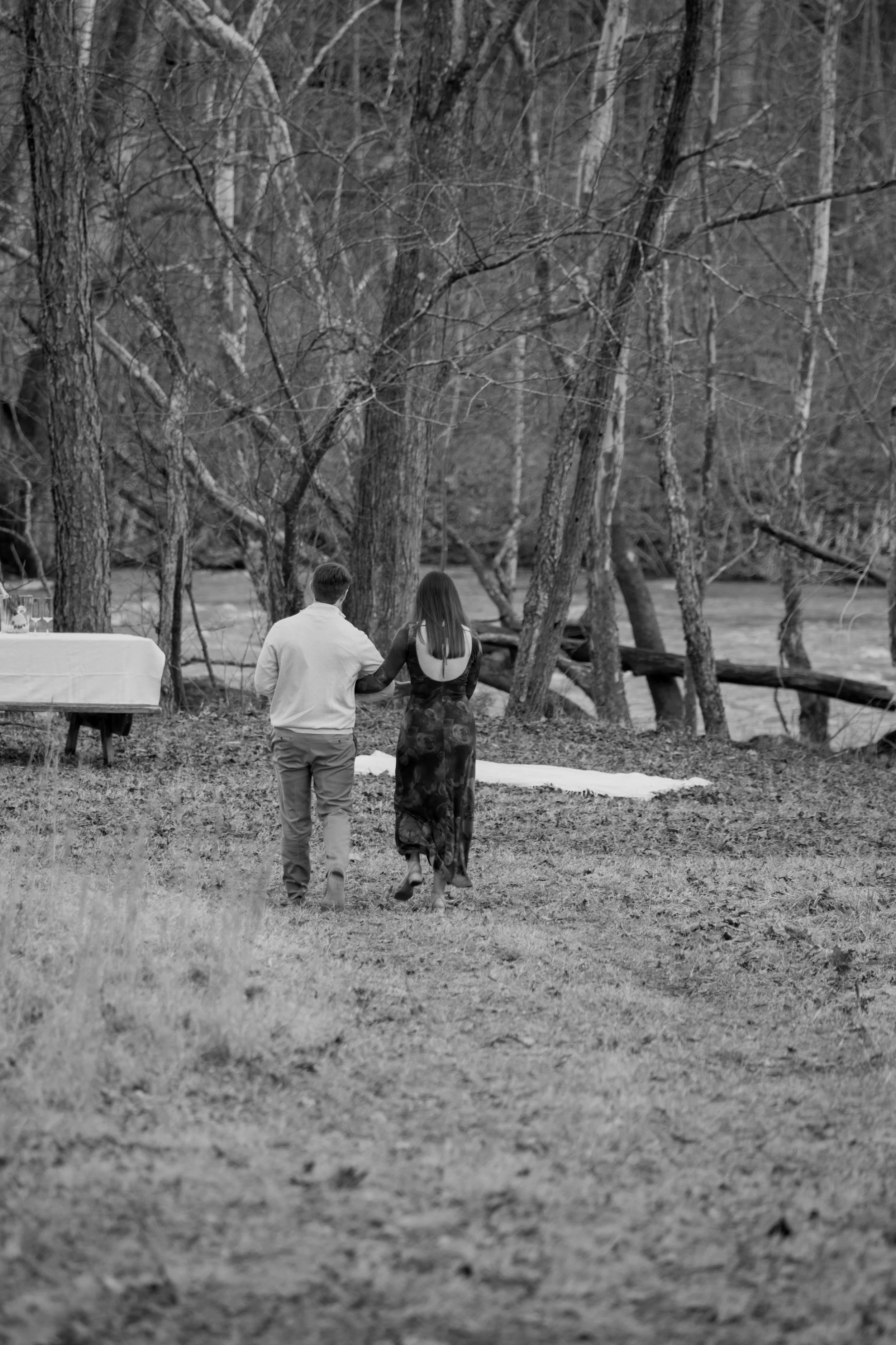 Engagement Photography by Will Locke near Richmond, VA in Montpelier. A couple walks on a white cloth on the ground near a wooded area with a stream on a fall day, with a table on the left side.