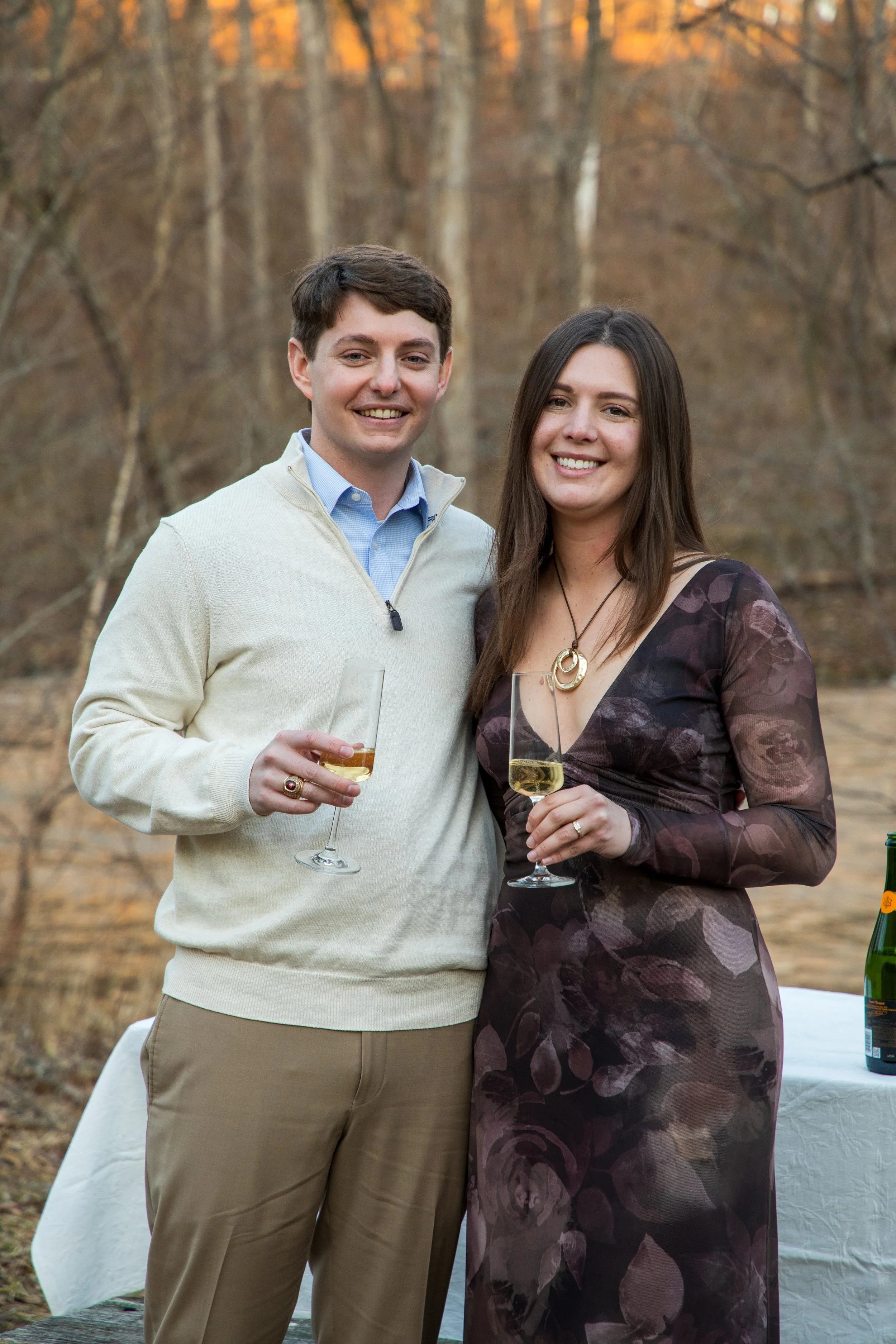 Engagement Photography by Will Locke near Richmond, VA in Montpelier. A man and woman smiling and hugging while holding champagne glasses outdoors during sunset, with a table of flowers nearby.