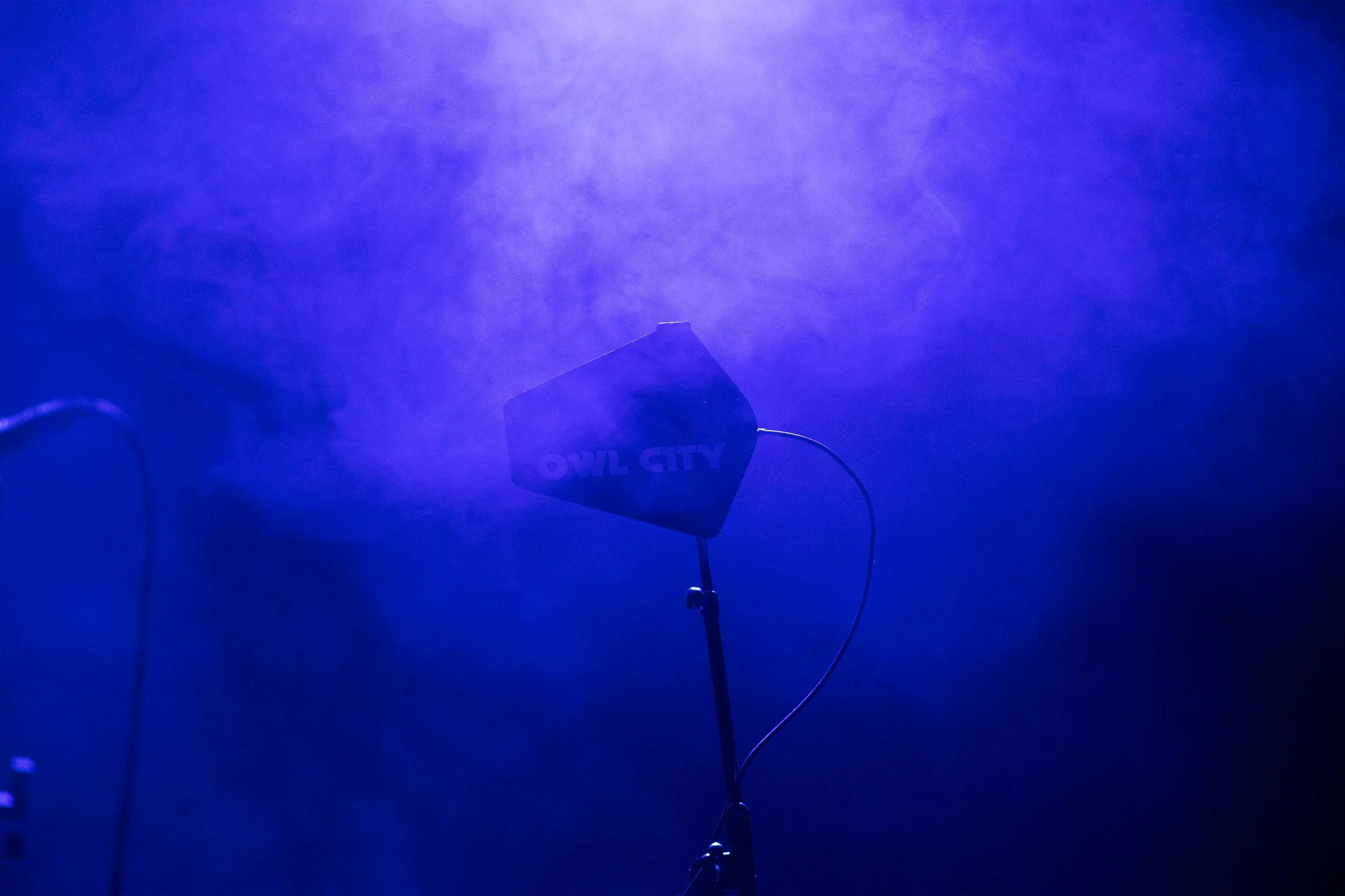 Concert Photography by Will Locke. A foggy scene with a spotlight shining down on a sign that reads 'OWL CITY', mounted on a stand with a wire attached. The background is dark with blue and purple hues.