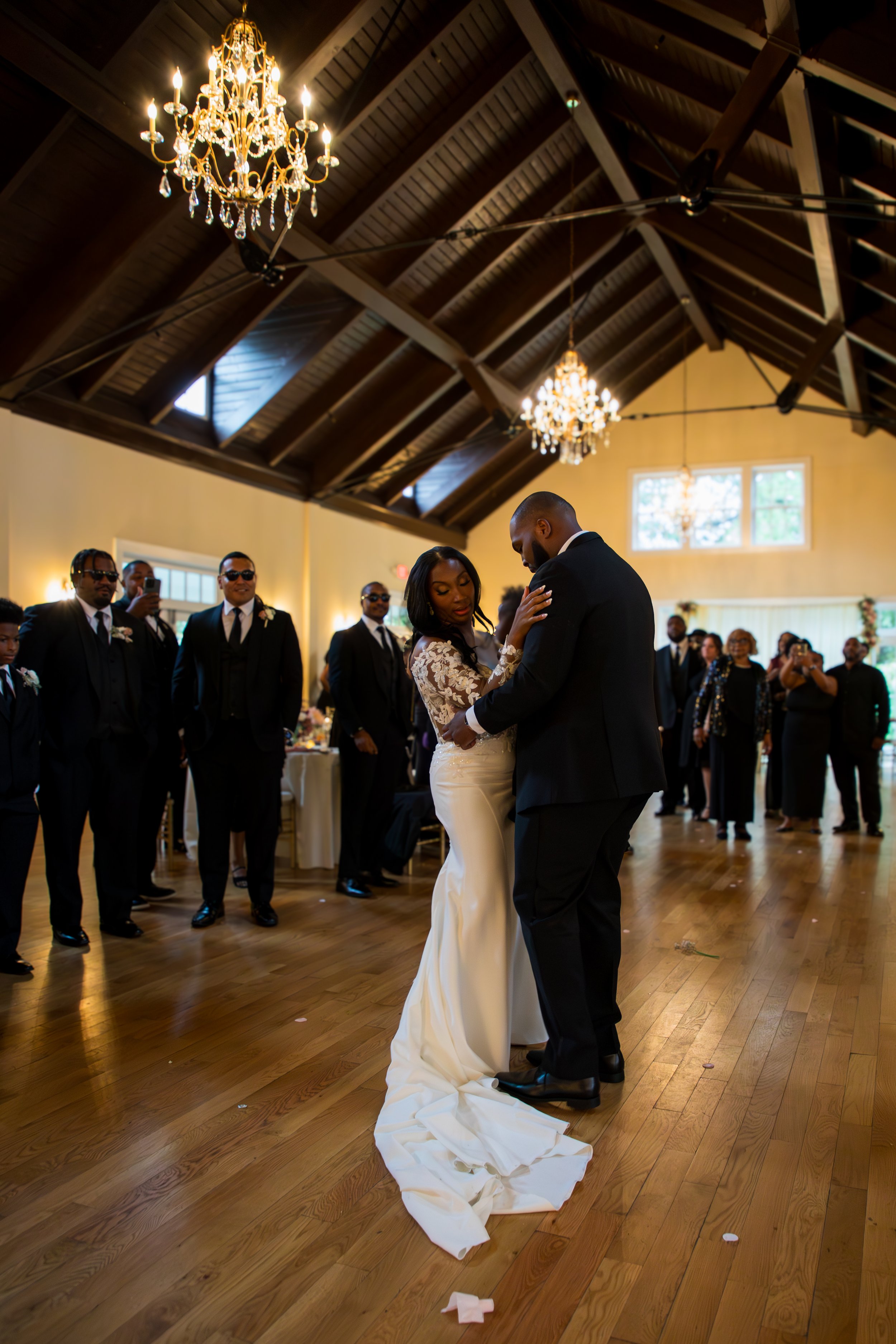 Photo & Video by Will Locke Wedding Photography at the Woman's Club of Portsmouth. Bride and groom share a dance at their wedding reception, surrounded by guests in formal attire, inside a decorated hall with chandeliers and wooden ceiling beams.