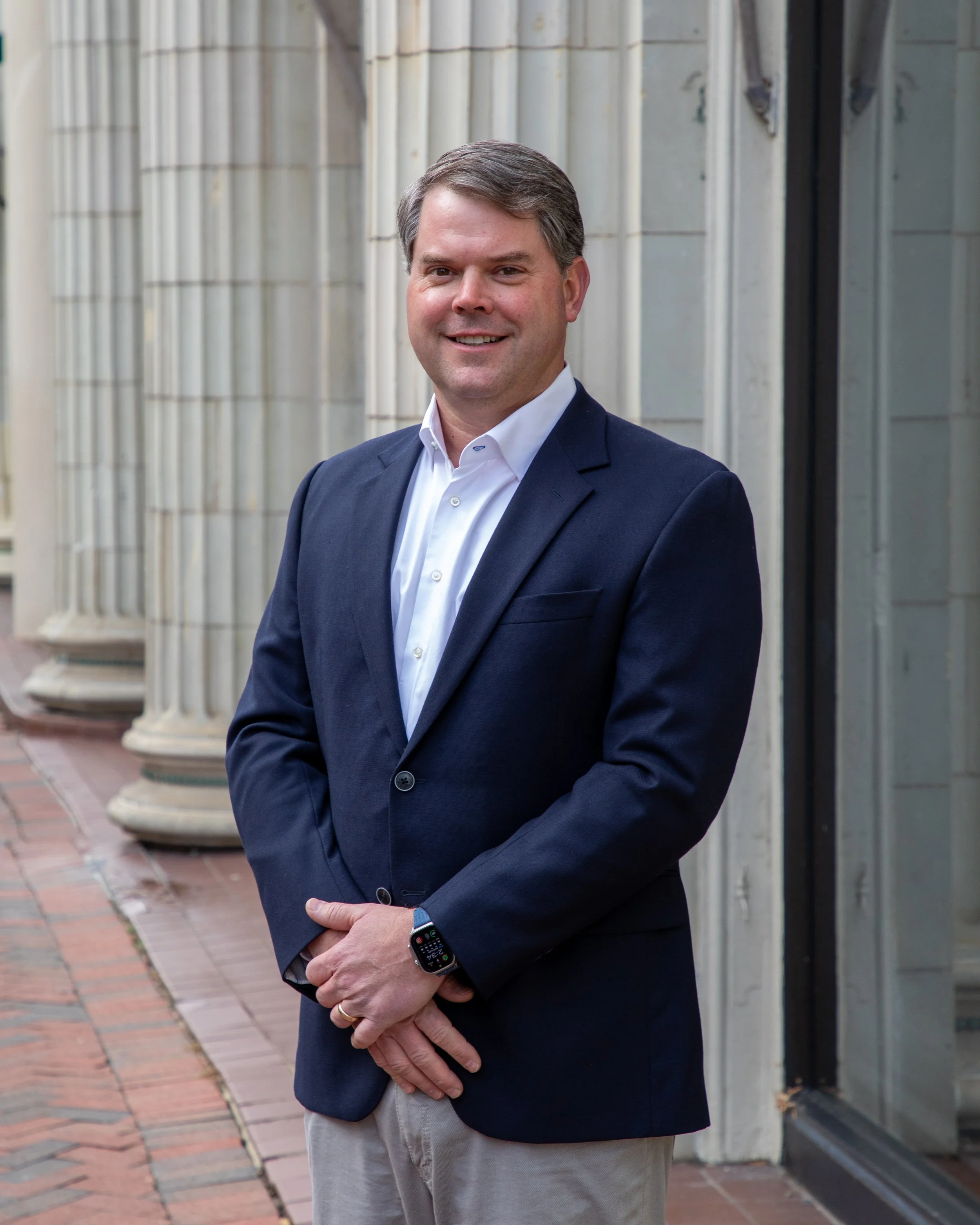Commercial Headshot Photography by Will Locke. A man in a navy blazer and white shirt standing outdoors in front of a row of columns.