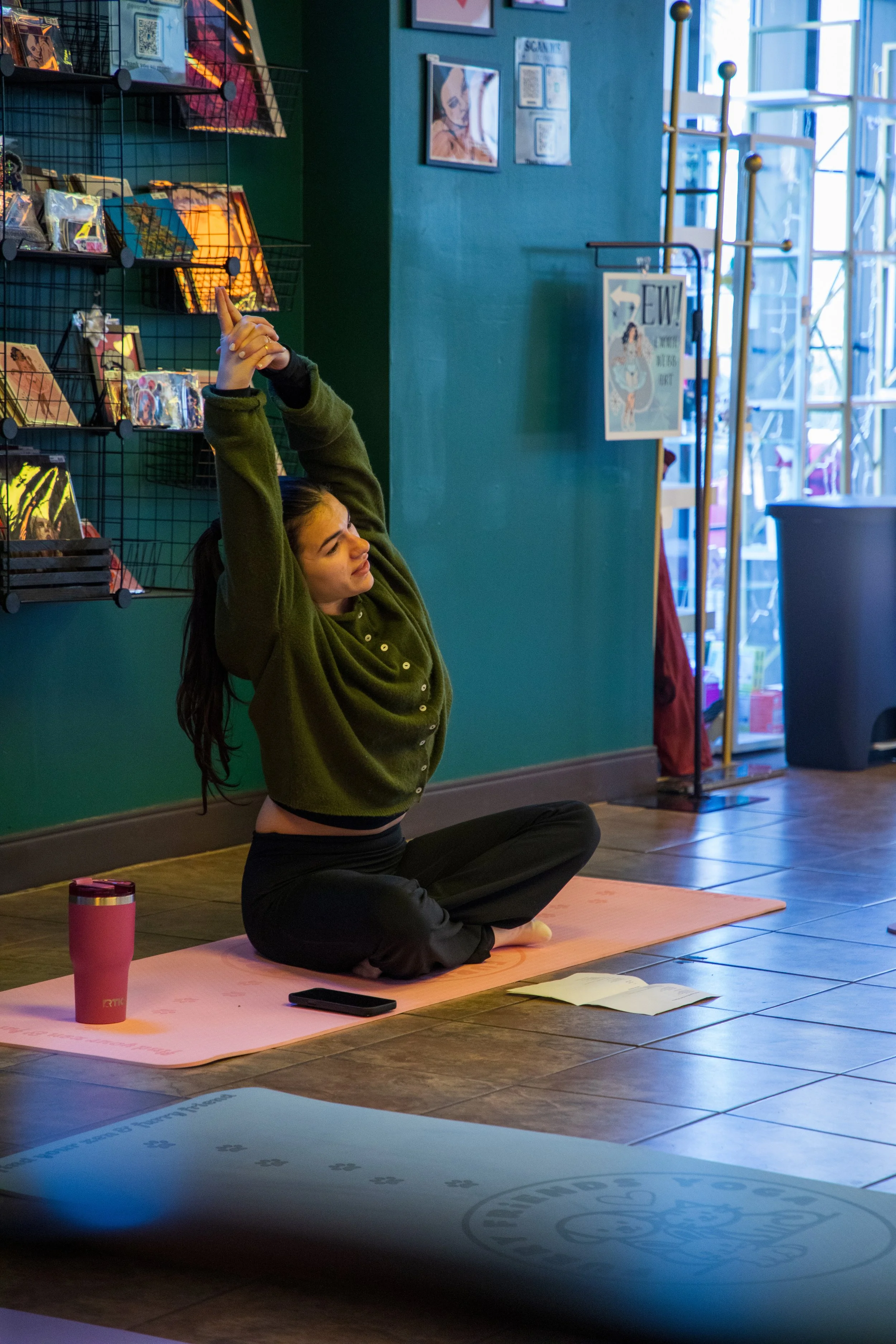 A woman sitting on a pink yoga mat, stretching her arms overhead, in a room with teal walls and large windows. Marketing and Event Photography by Will Locke. 