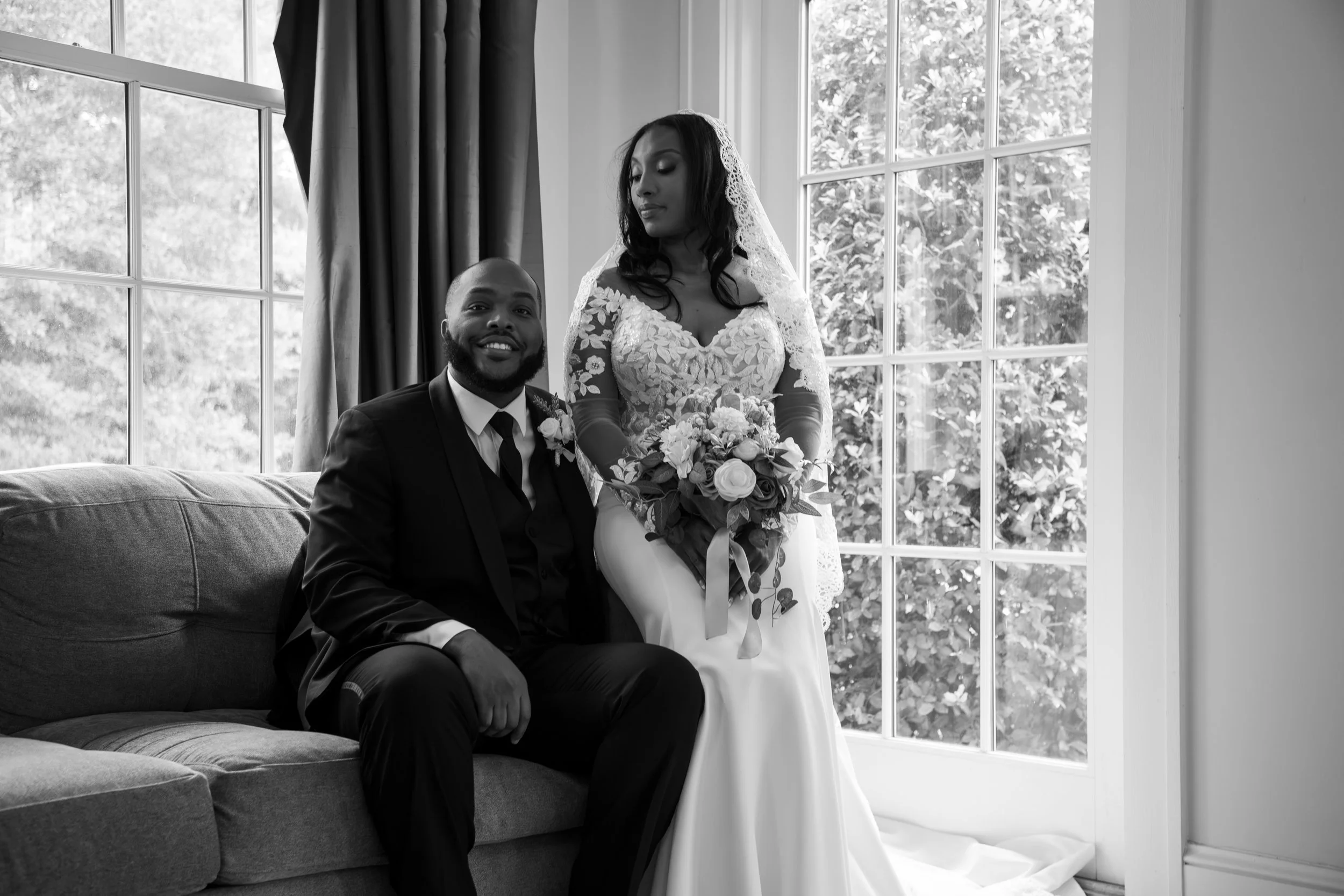Photo & Video by Will Locke Wedding Photography at the Woman's Club of Portsmouth in VA. A bride and groom in wedding attire sitting on a sofa by a large window, with the bride holding a bouquet of flowers.