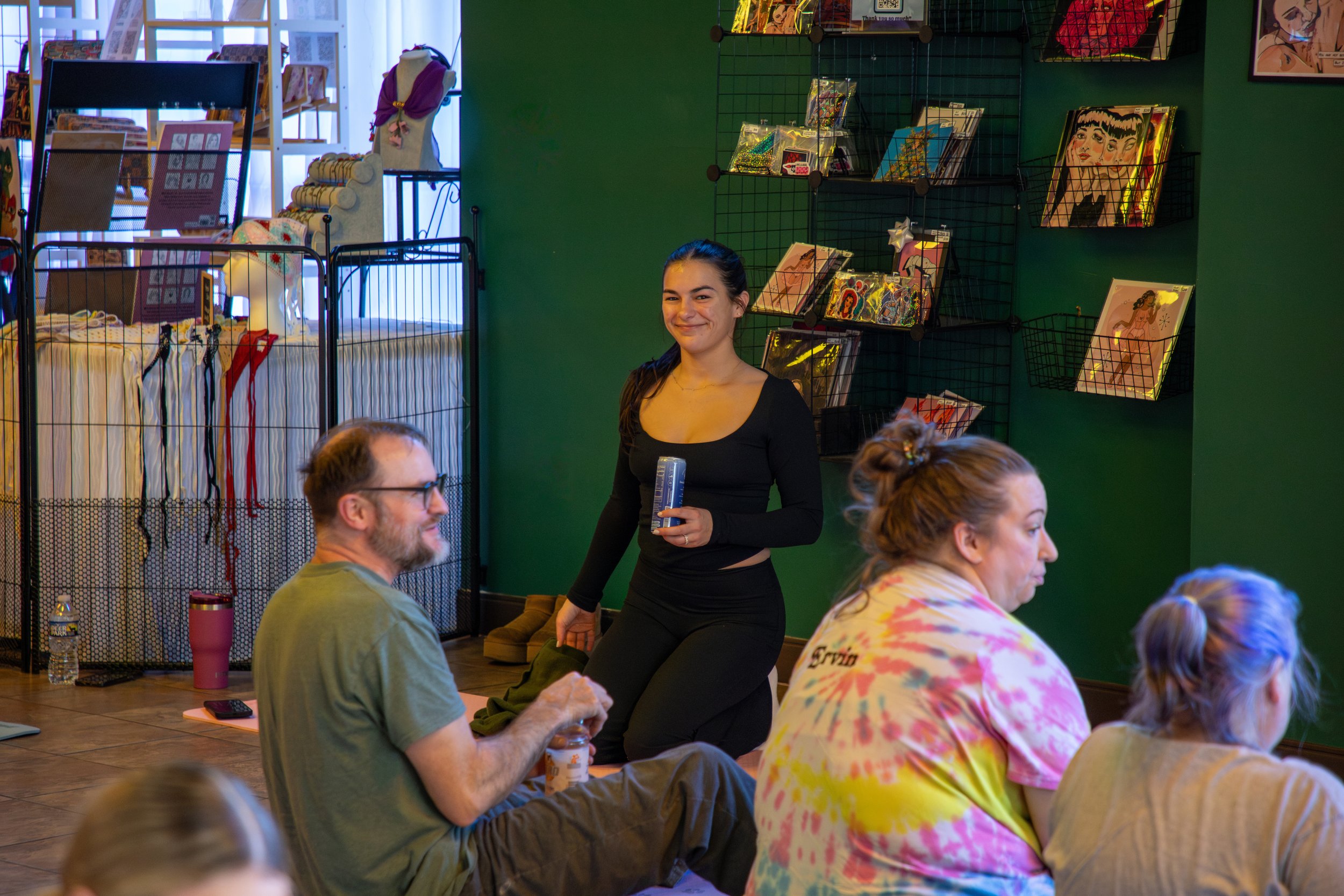Marketing and Event Photography by Will Locke. A young woman with dark hair smiling and holding a box, standing behind three seated people in a colorful, artsy room with green walls and wall-mounted magazine racks.