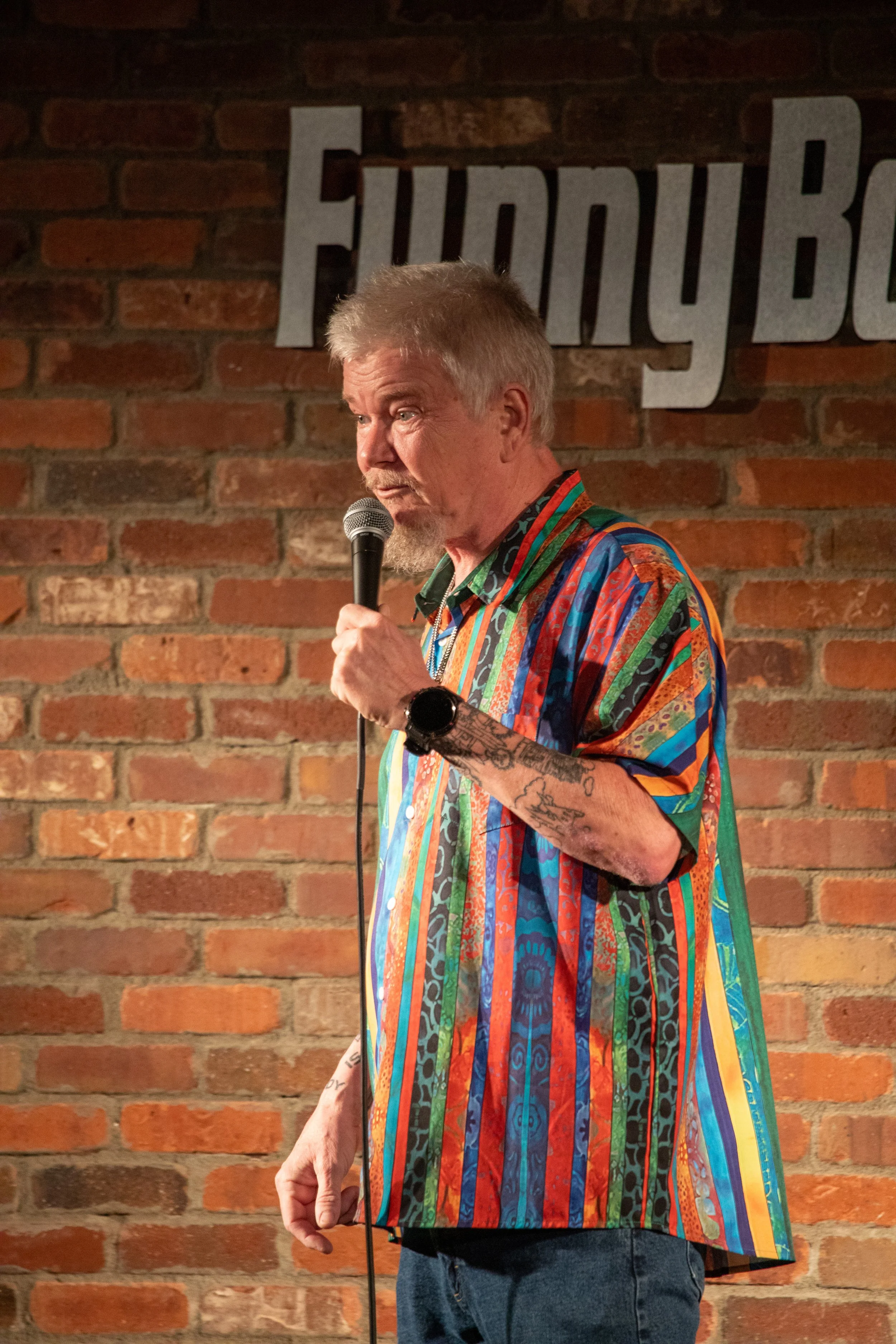 Comedy Photography by Will Locke. A man with gray hair, tattoos, and a beard performs stand-up comedy on stage, holding a microphone, in front of a brick wall and white block letters spelling "Funny Bone."