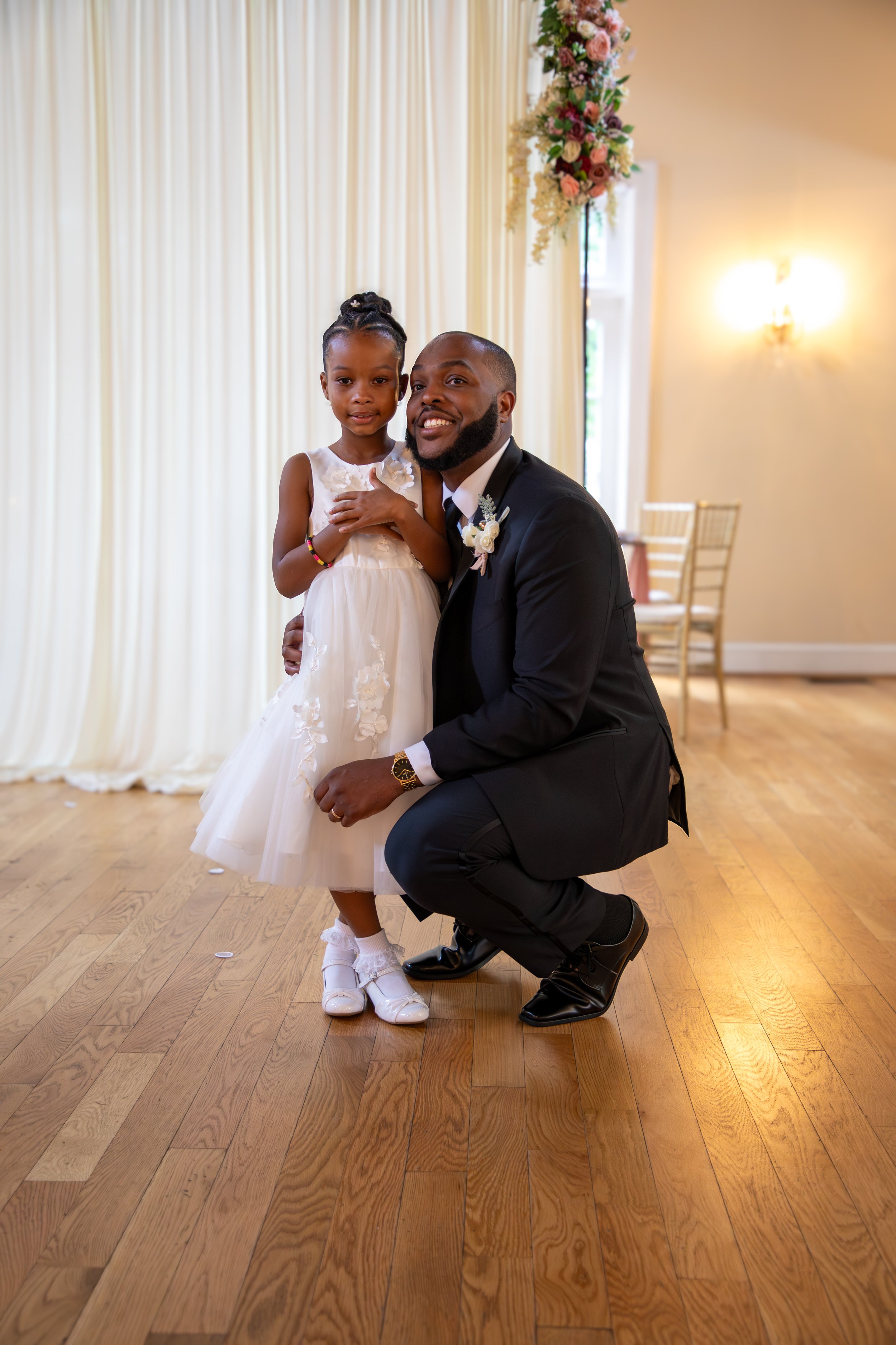 Photo & Video by Will Locke Wedding Photography at the Woman's Club of Portsmouth in Virginia. A man in a black suit kneeling next to a young girl in a white dress at a wedding reception or ceremony.