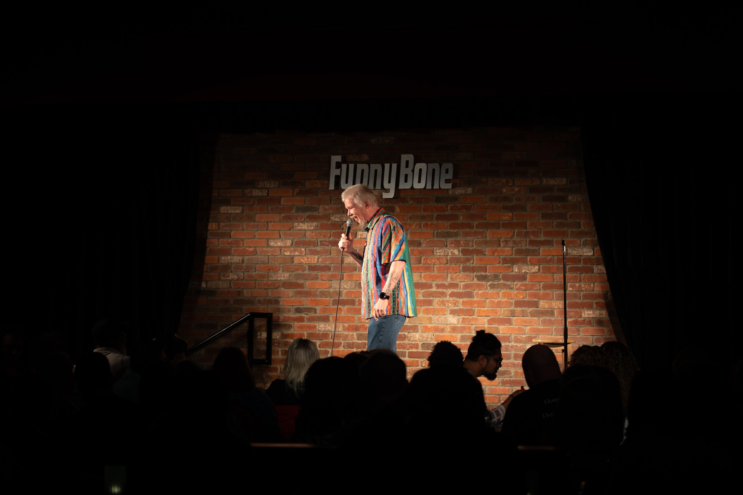 Comedy Photography by Will Locke. A comedian on stage at Funny Bone, holding a microphone and wearing a colorful, striped shirt, performing in front of a brick wall background with the Funny Bone sign.