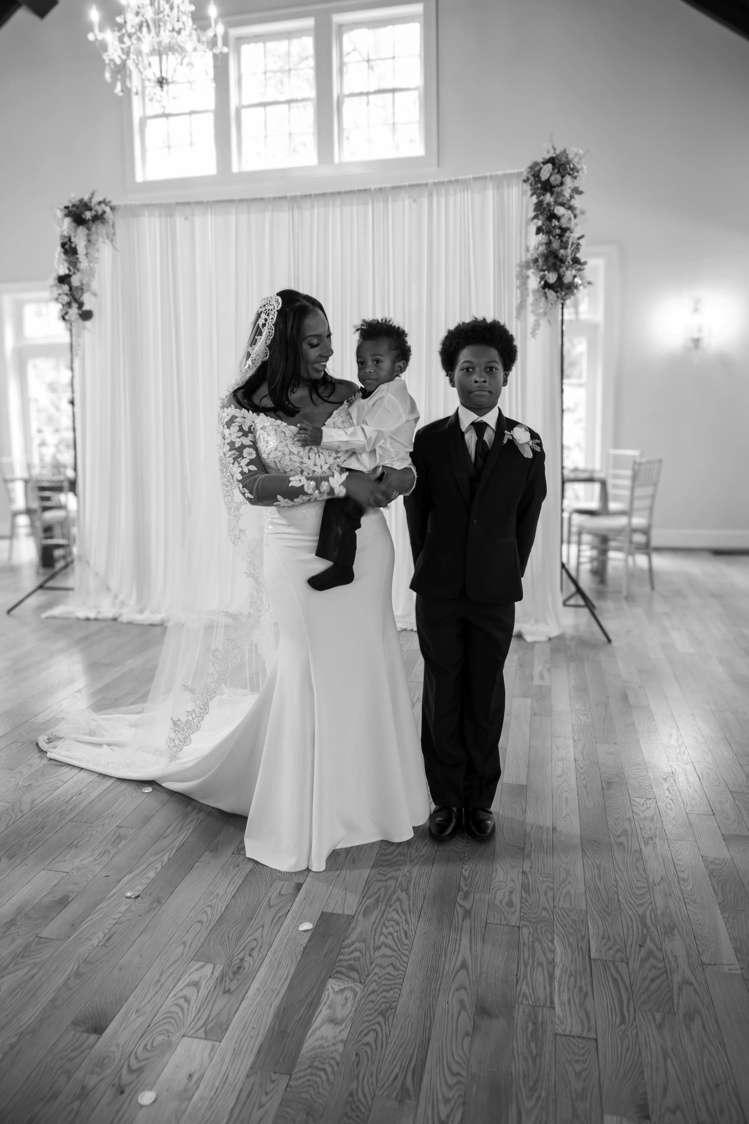 Photo & Video by Will Locke Wedding Photography at the Woman's Club of Portsmouth in Virginia. Black-and-white photo of a woman in a wedding dress holding a young child, standing next to a young man in a suit, in a decorated indoor venue.