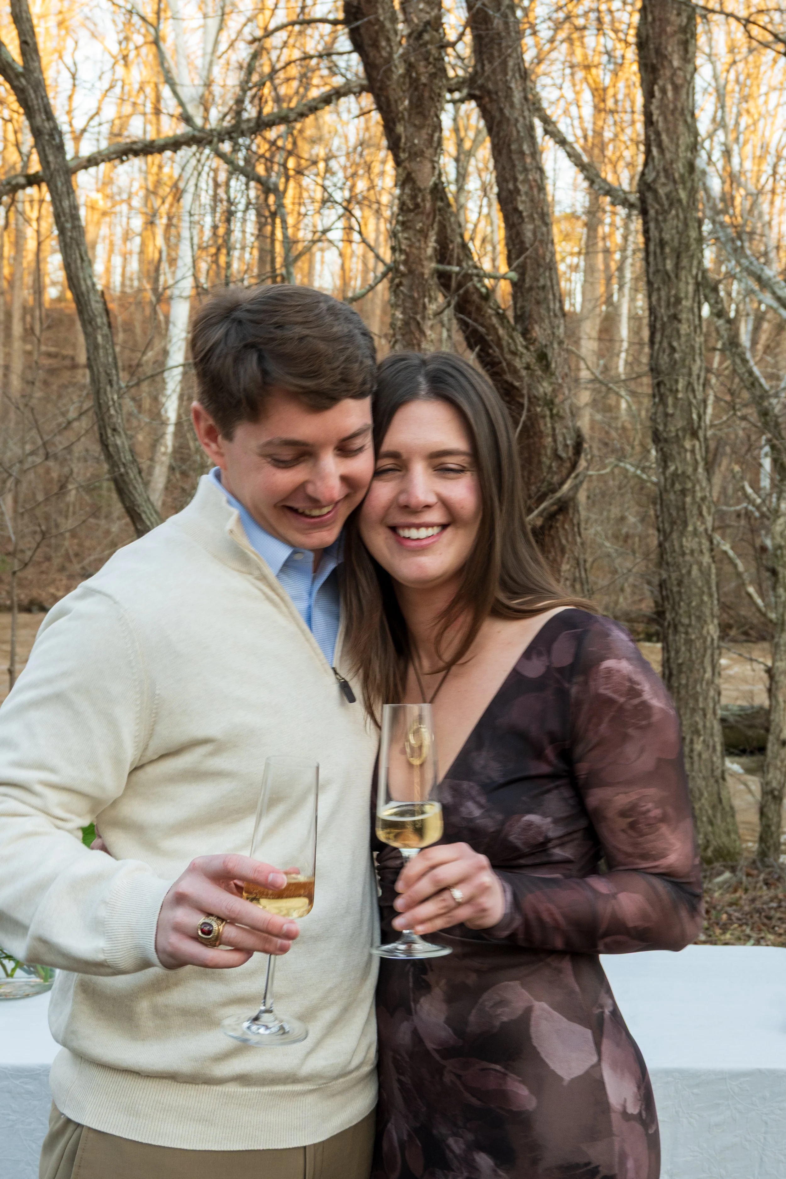Engagement Photography by Will Locke near Richmond, VA in Montpelier. A man and woman smiling and hugging while holding champagne glasses outdoors during sunset, with a table of flowers nearby.