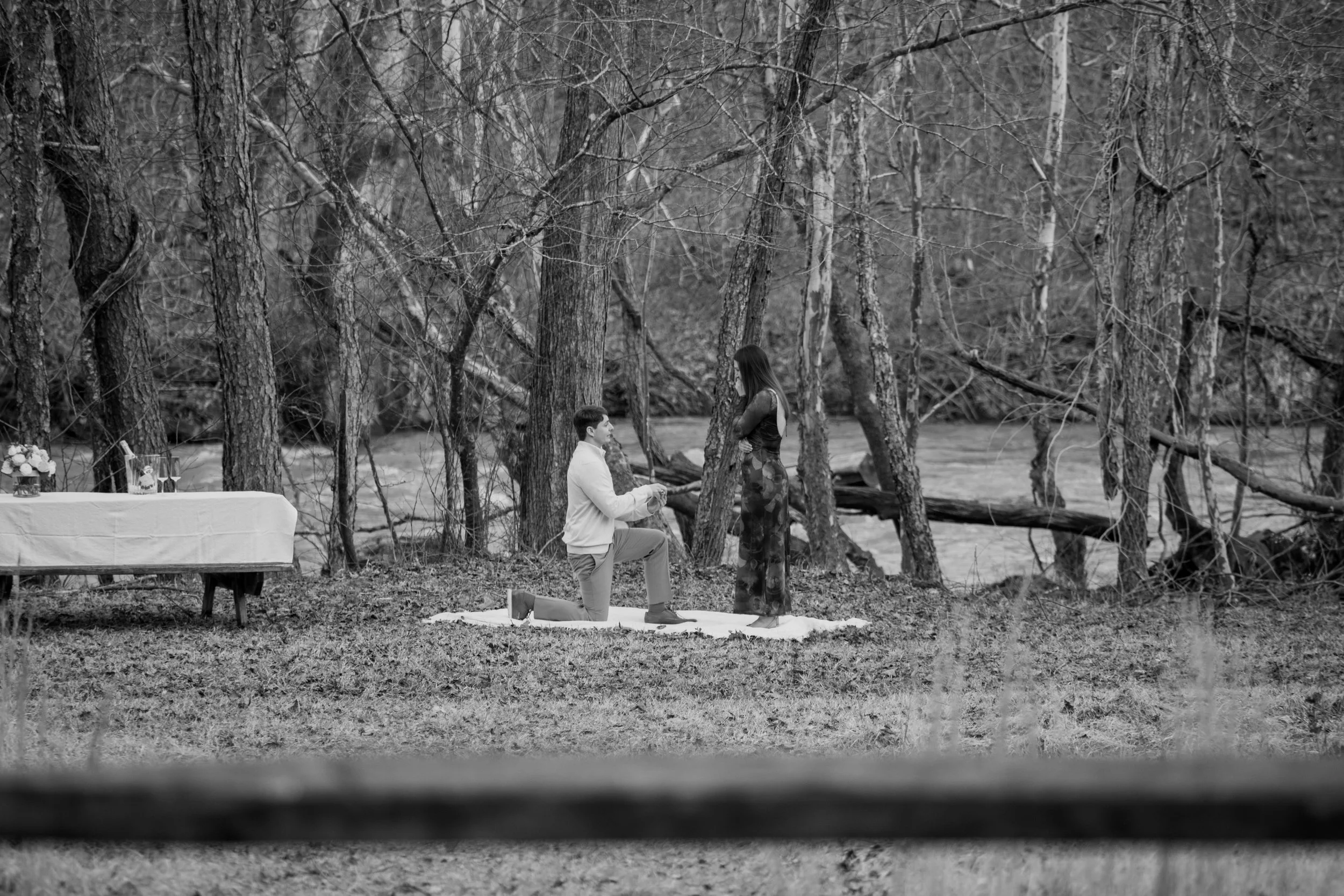 Engagement Photography by Will Locke near Richmond, VA in Montpelier. A man proposing to a woman outdoors in a wooded area near a river, with a small table set with drinks nearby.