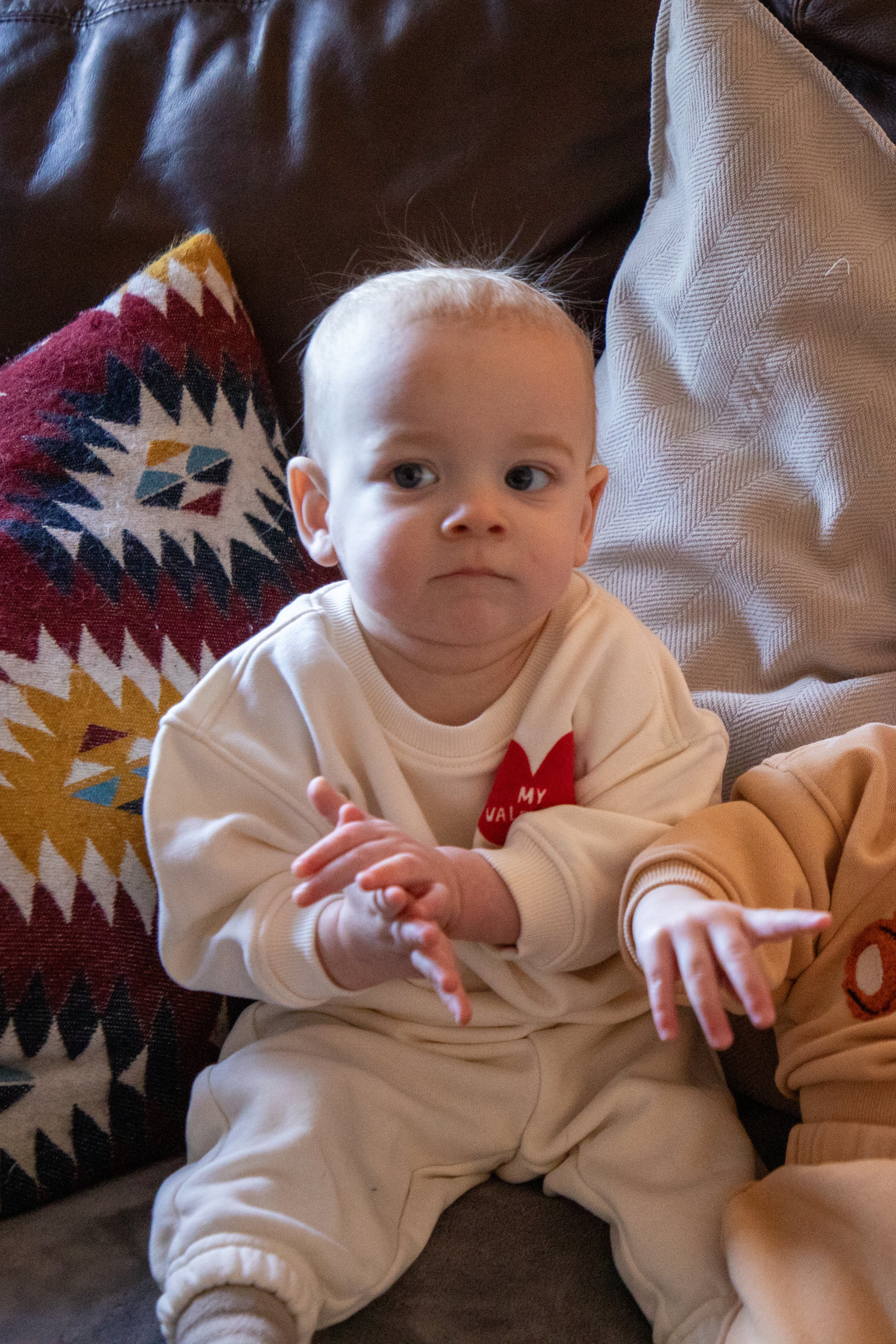 Newborn Baby Photography by Will Locke. A young child with light-colored hair sitting on a couch, looking to the side with a neutral expression, wearing a white sweatshirt with a red heart design and cream pants.