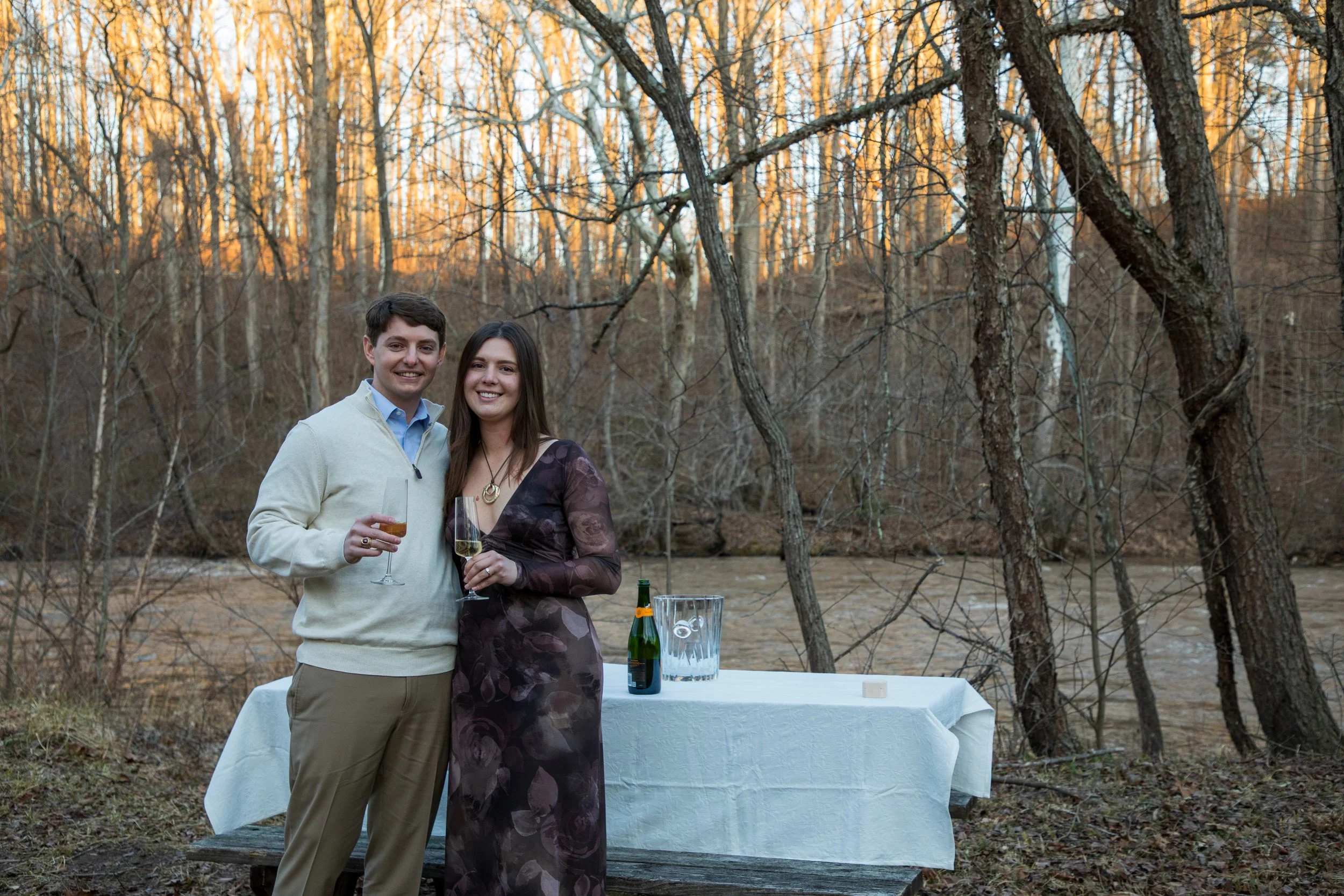 Engagement Photography by Will Locke near Richmond, VA in Montpelier. A couple standing outdoors by a table with drinks, in a wooded area during sunset.