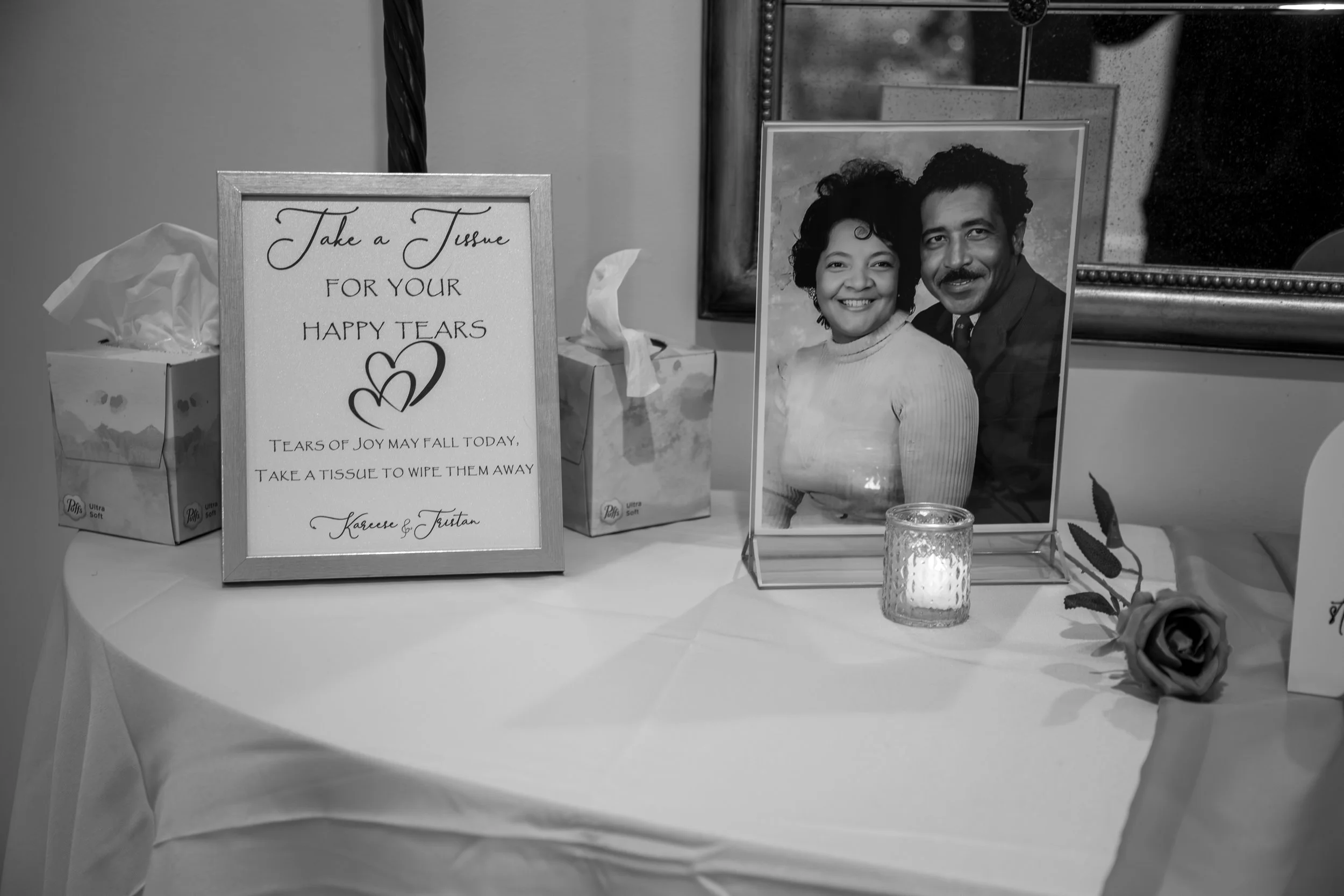 Wedding Photography by Will Locke. A tribute display with a framed sign, a framed photo of a smiling couple, a candle, and a fake rose on a table. The sign reads 'Take a Tissue for Your Happy Tears. Tears of joy may fall today.