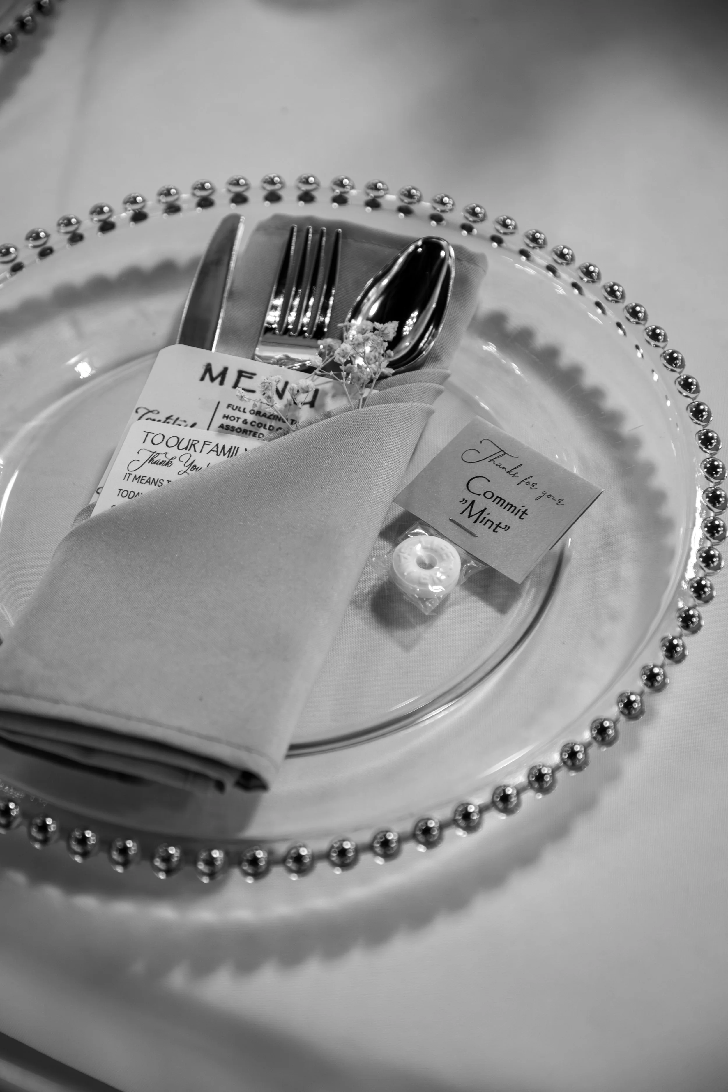 Wedding Photography by Will Locke. Elegant table setting with a clear plate bordered by silver beads, silverware wrapped in a napkin, a small flower, a thank you note, and a mint on a white tablecloth.