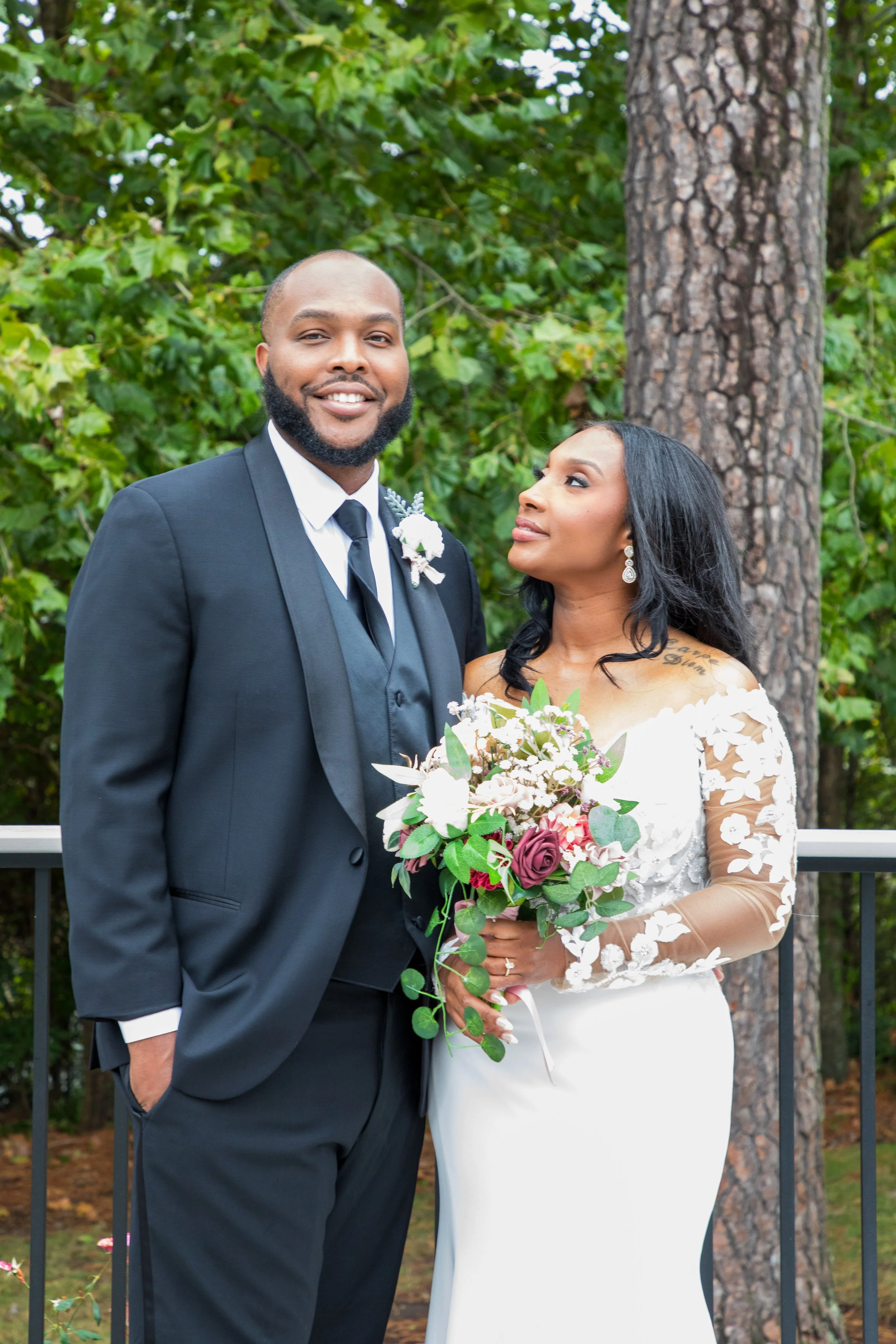 Photo & Video by Will Locke Wedding Photography at the Woman's Club of Portsmouth in VA. A newlywed couple posing outdoors in front of trees, the man in a black tuxedo and the woman in a white wedding dress holding a bouquet of flowers.