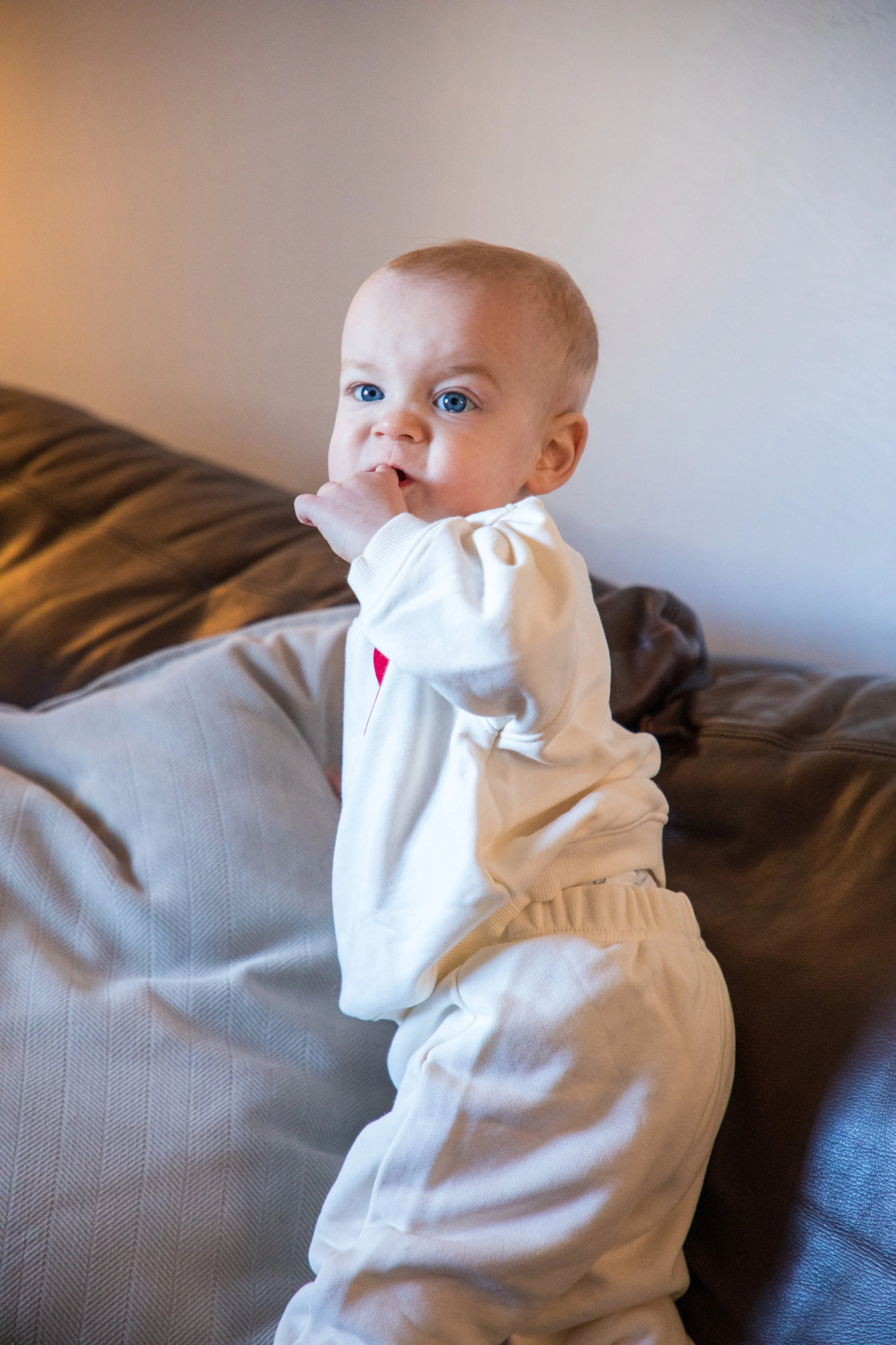 Baby Photography by Will Locke. A young child with blue eyes and light hair standing on a couch, with one hand in their mouth, wearing cream-colored pajamas.